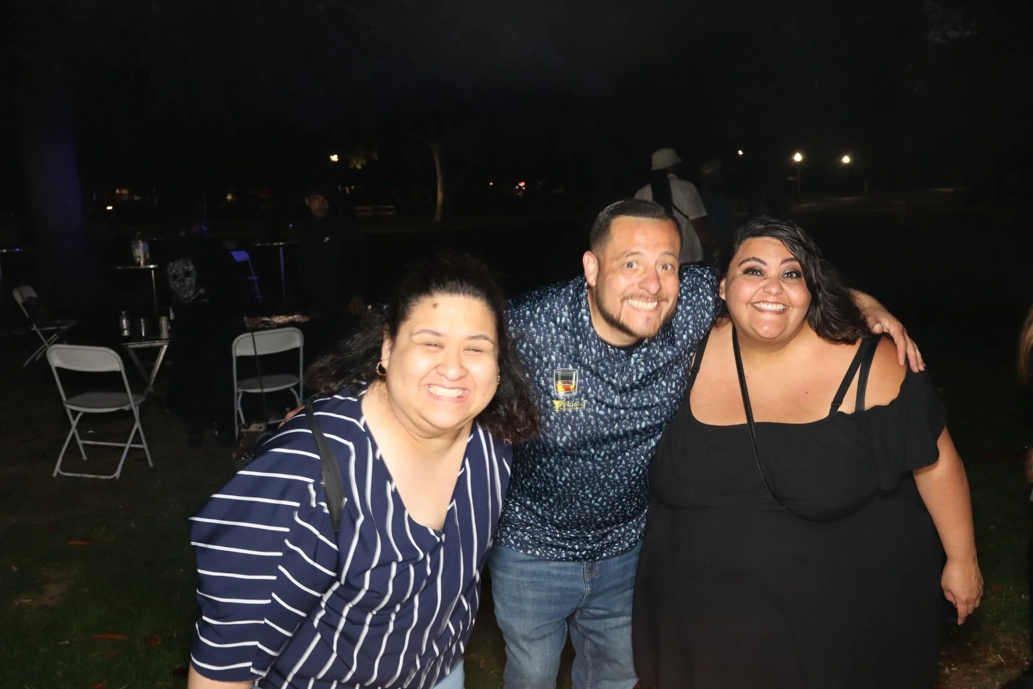 Group of three people smiling at night outdoors, with chairs and trees in the background.