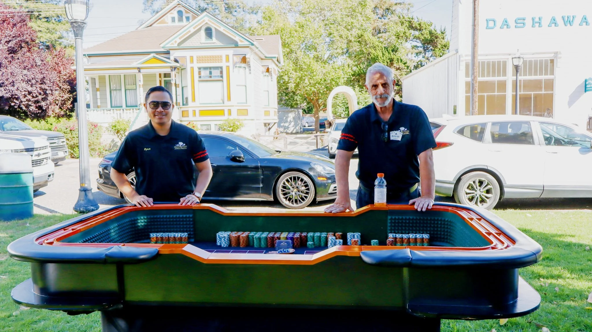 Two men standing behind a casino poker table outdoors, with parked cars and a house in the background. One man is wearing sunglasses, and both are dressed in black shirts with logos.