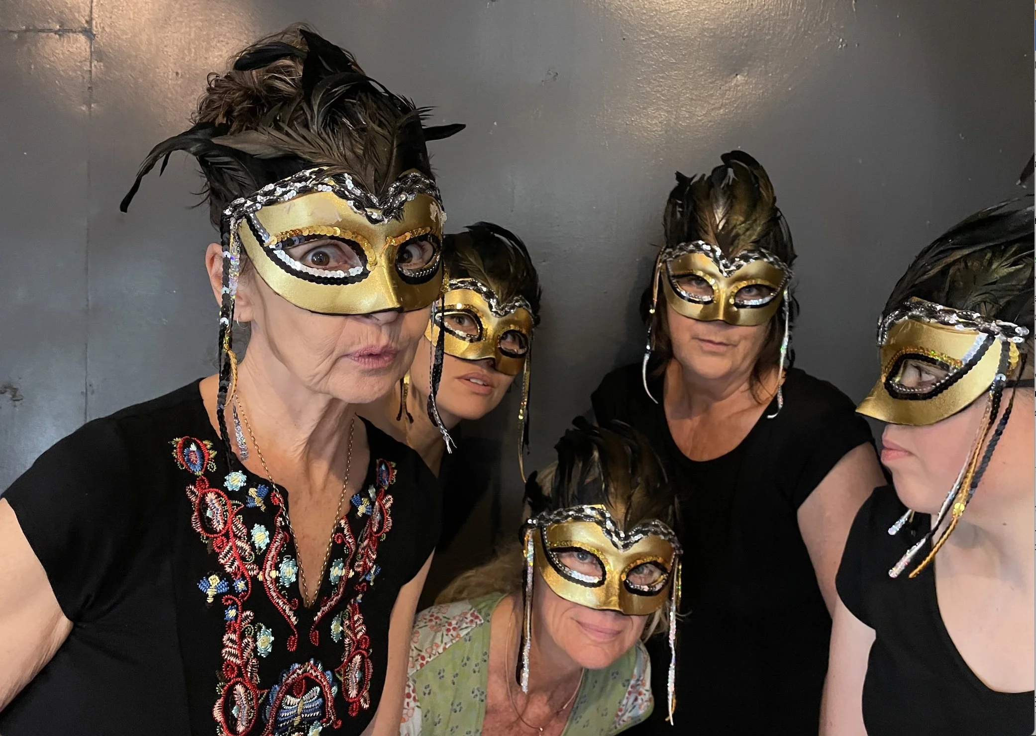 Five women wearing gold, black, and sequin decorated masks with feathers, standing together against a dark wall.