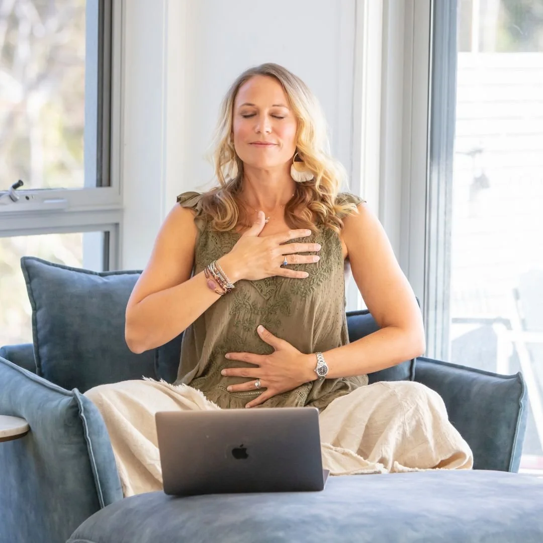 Founder, Ann Marie, sitting cross-legged on a couch, with hands on chest and abdomen, eyes closed, in front of a laptop.