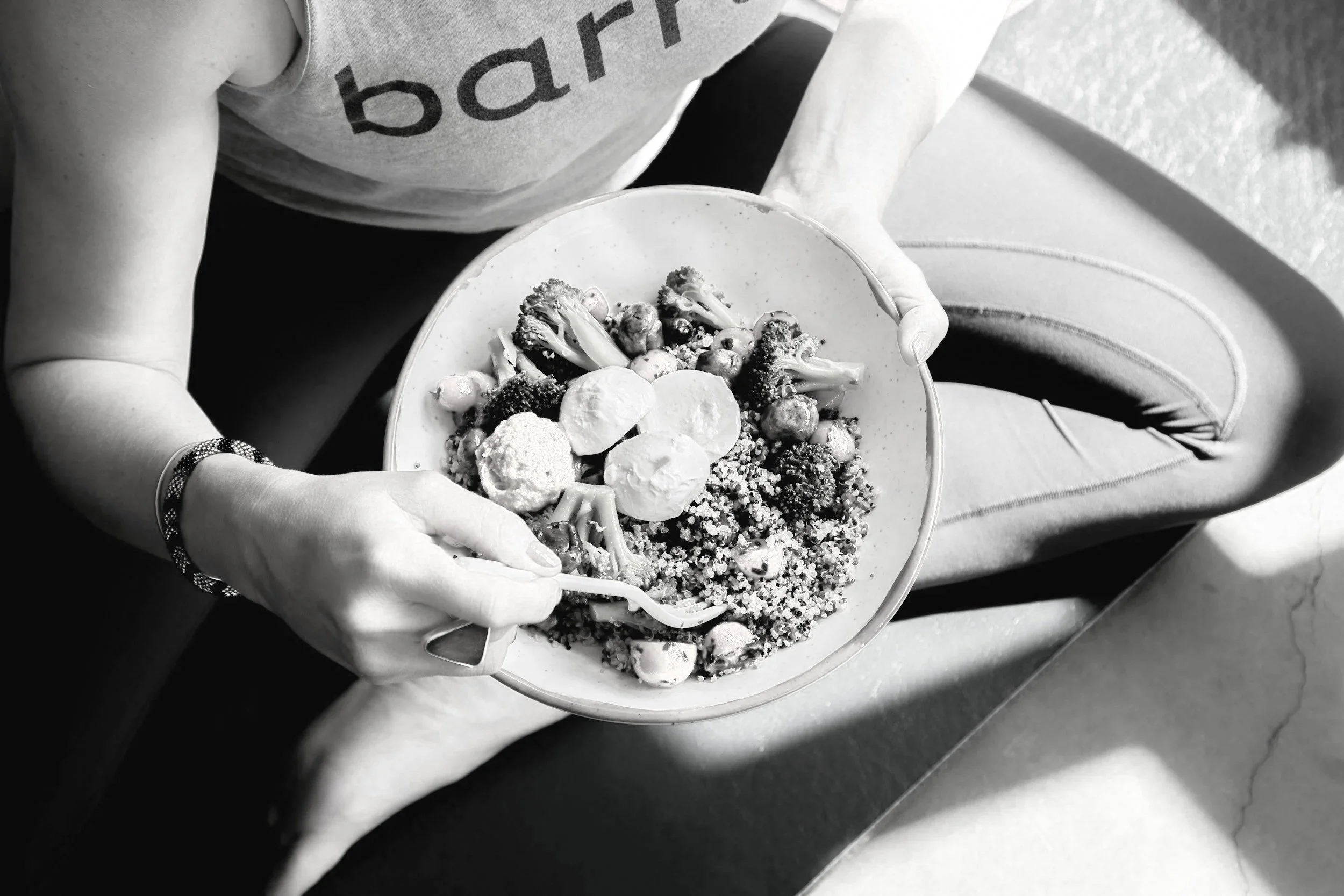 A person holding a bowl of salad and sitting on a chair, wearing a tank top with the word 'barr' visible, with a bracelet on their left wrist, and a fork in their right hand.