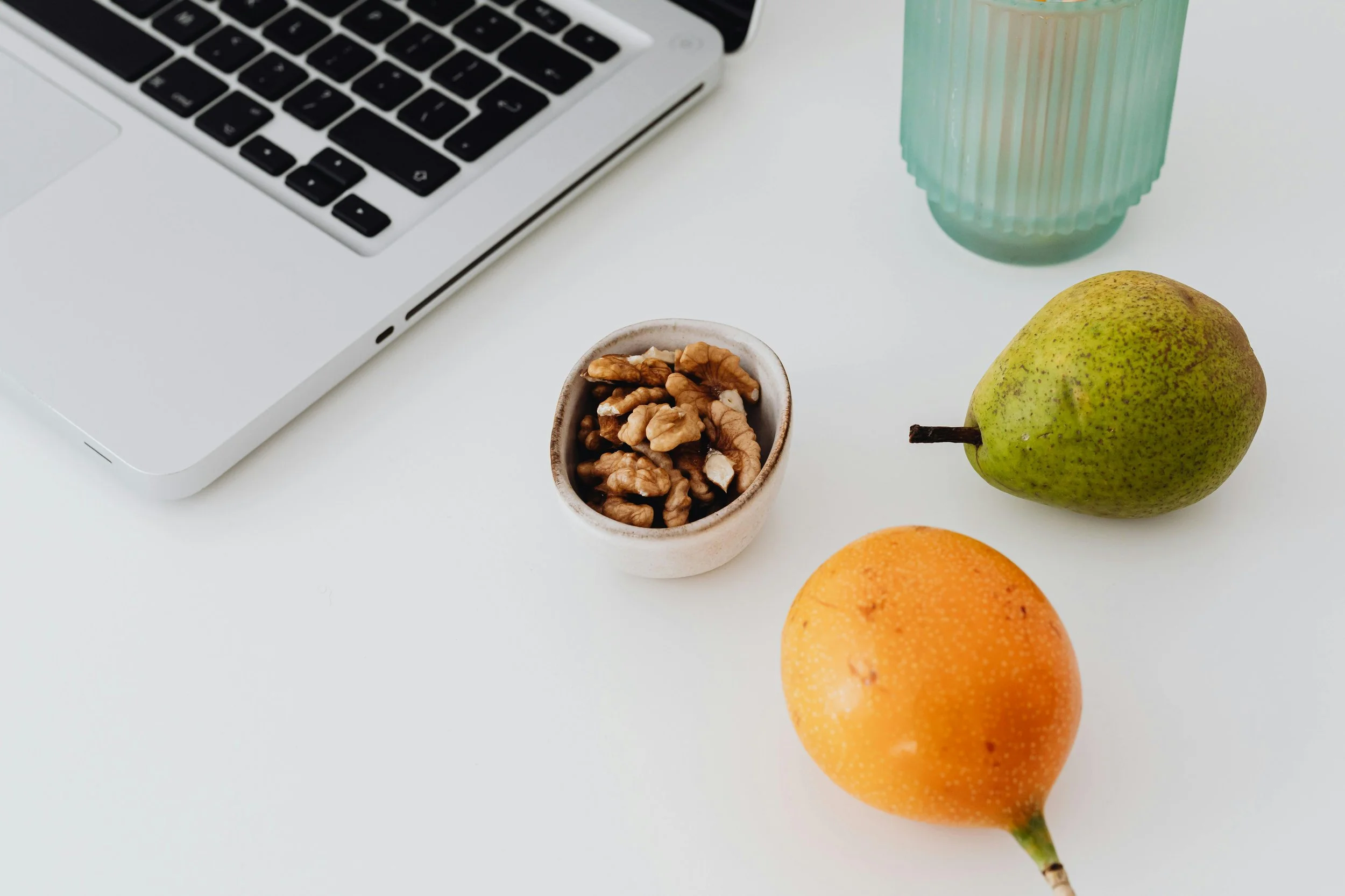 A white desk with a laptop, a small bowl of walnuts, a green pear, an orange, and a tall, green ribbed glass of water.