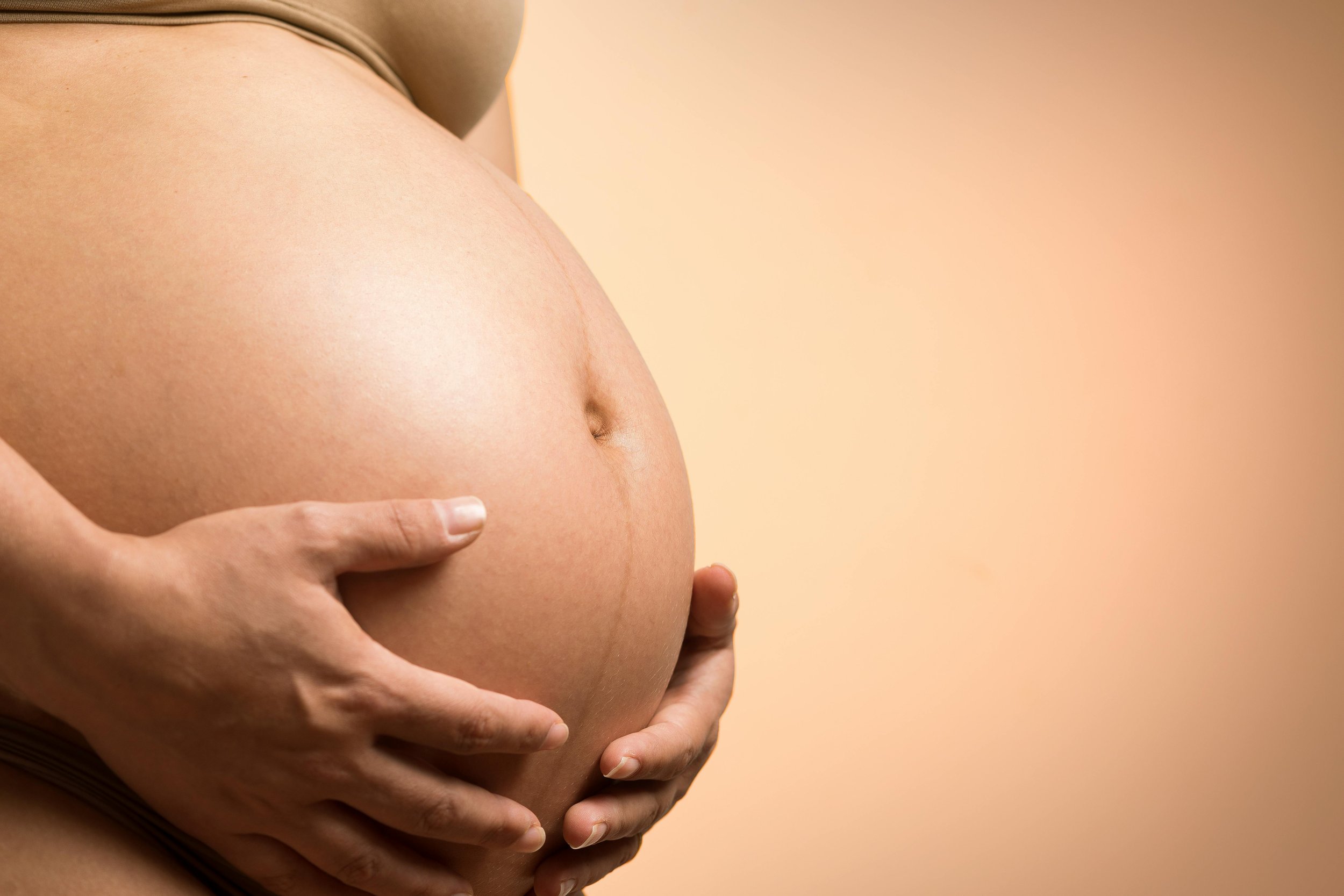 Close-up of a pregnant woman's belly being gently held with both hands, with a soft peach background.