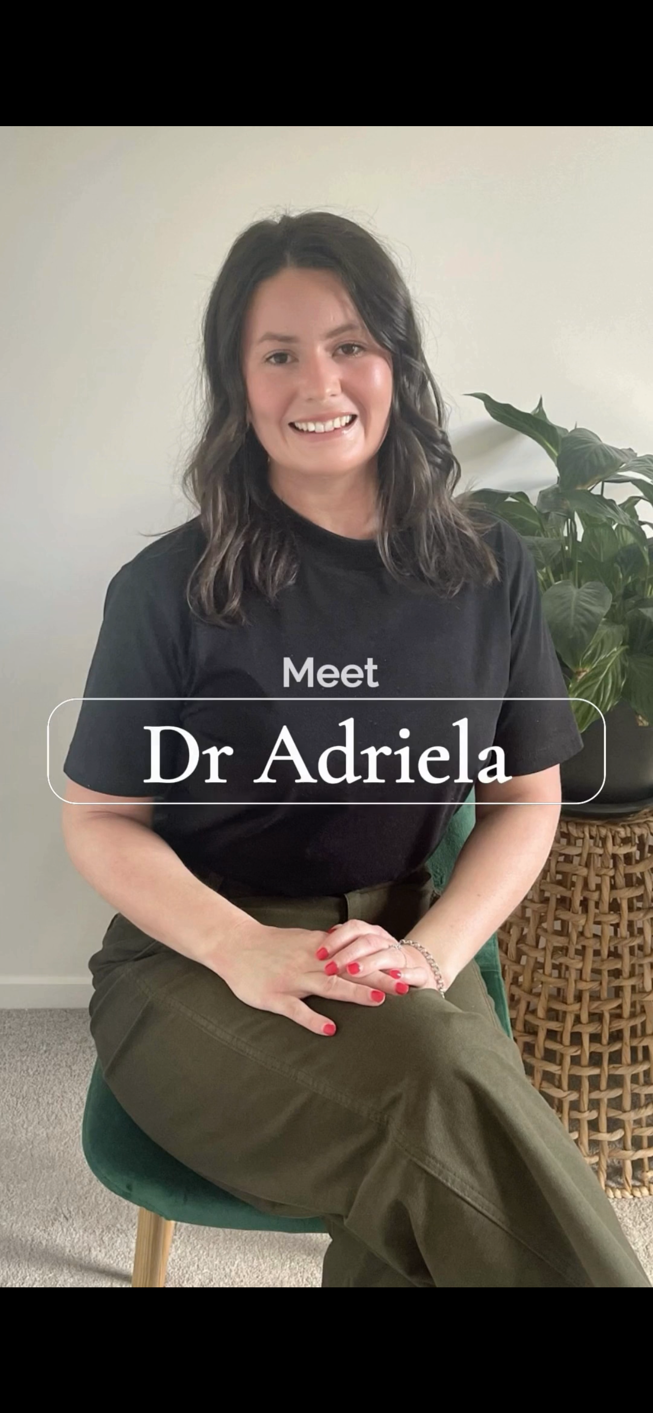 A woman with dark, wavy hair sitting on a green chair, smiling at the camera. She is wearing a black T-shirt and olive green pants, with her hands folded in her lap. There is a large leafy plant behind her and a woven basket beside her.