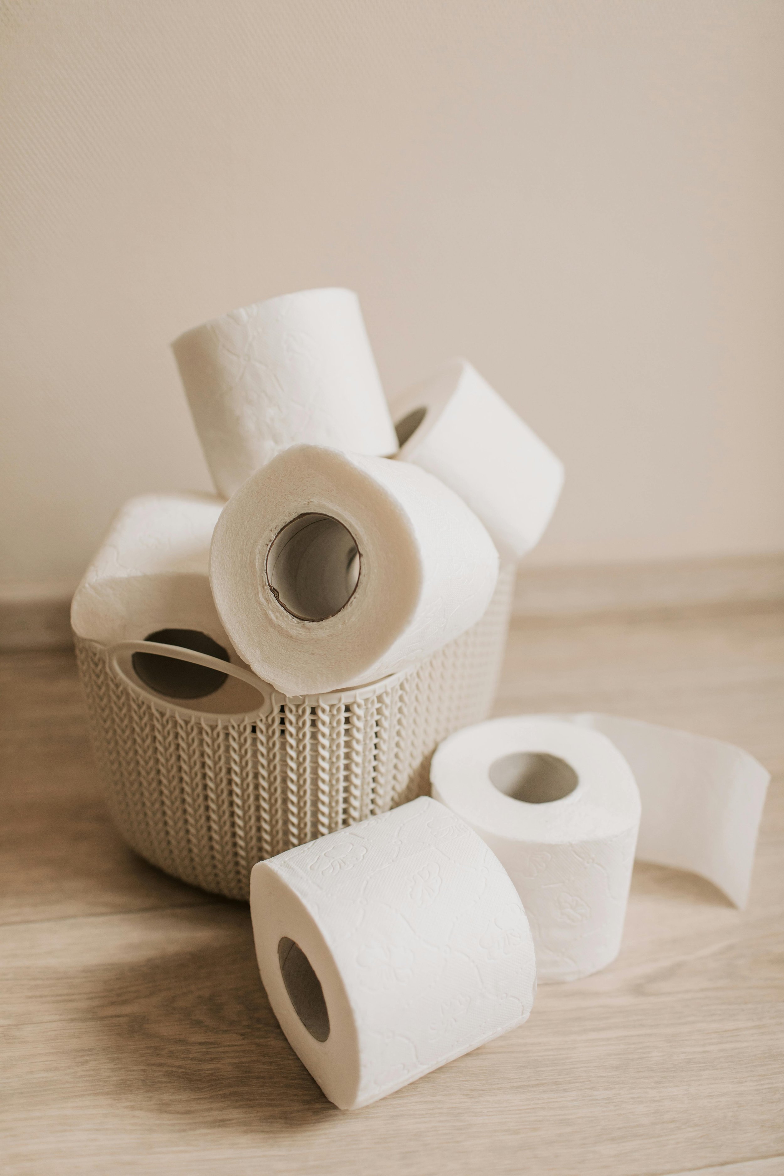 A basket filled with several rolls of white toilet paper, some of which are placed on a wooden surface in front of the basket.