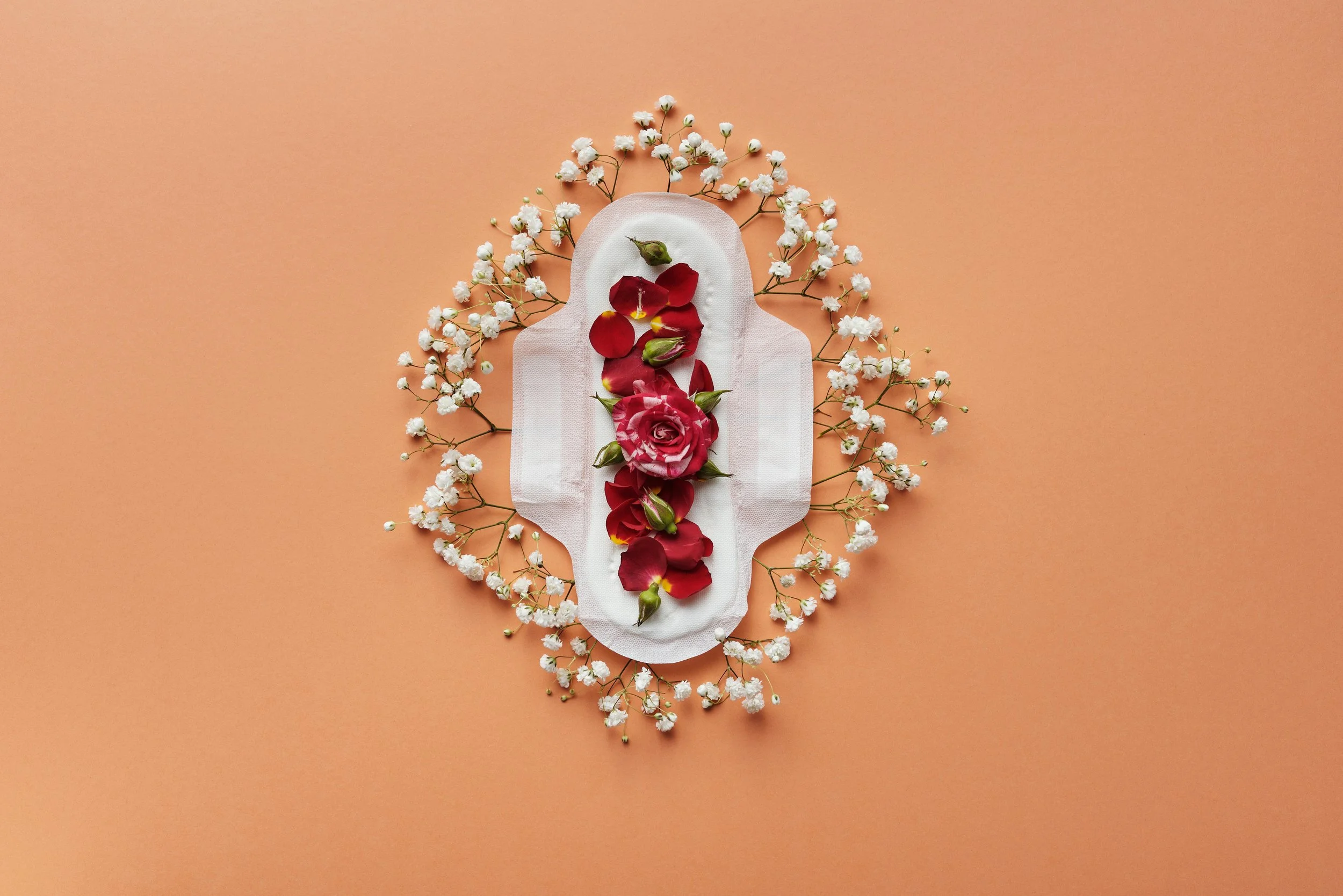 A sanitary pad decorated with red and pink rose petals and surrounded by baby's breath flowers on a peach background.