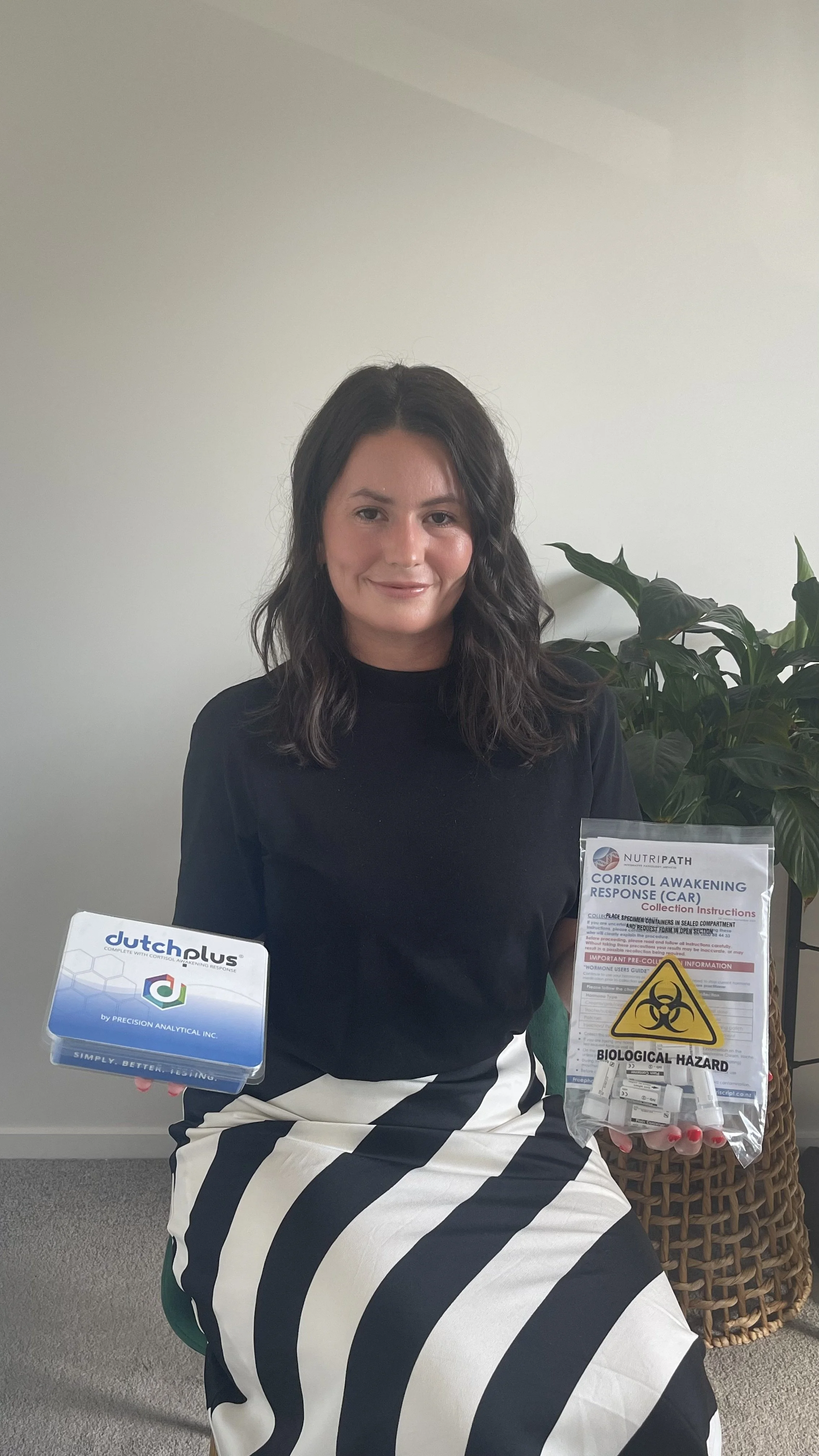 Dr Adriela from 'Future You Wellness' - 'Functional Medicine' sitting indoors, holding a box labeled 'Dutch Plus' in one hand and a sealed bag with biohazard warning and medical supplies in the other, next to a large green plant.