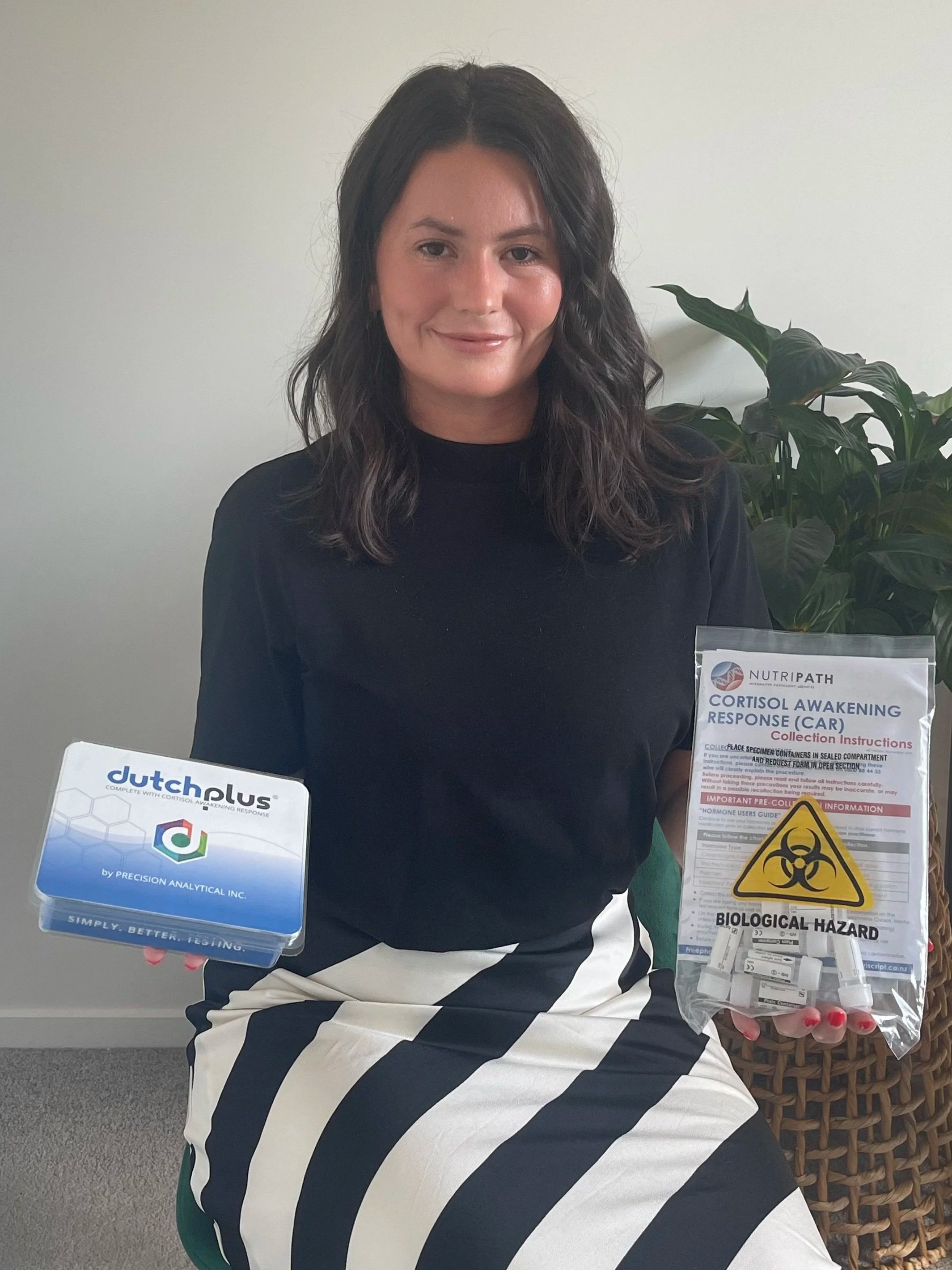Dr Adriela from 'Future You Wellness' - 'Functional Medicine' sitting indoors, holding a box labeled 'Dutch Plus' in one hand and a sealed bag with biohazard warning and medical supplies in the other, next to a large green plant.