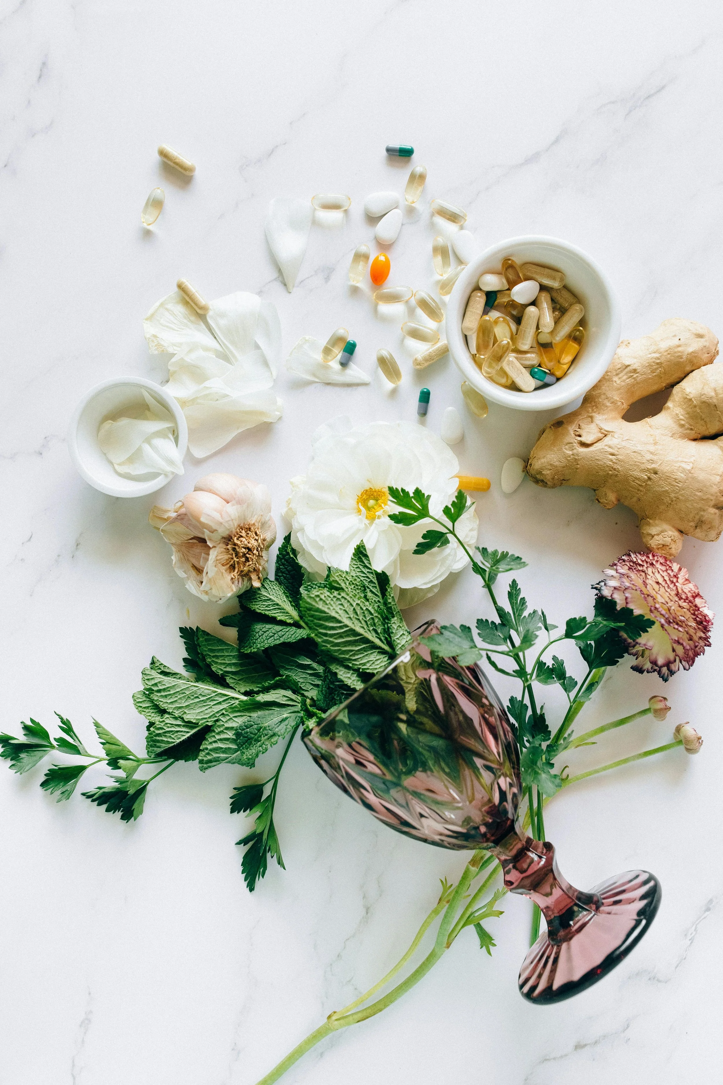 A flat lay of various pills, tablets, and capsules in bowls and scattered on a white marble surface, alongside fresh herbs, flowers, a piece of ginger, and a pink glass.