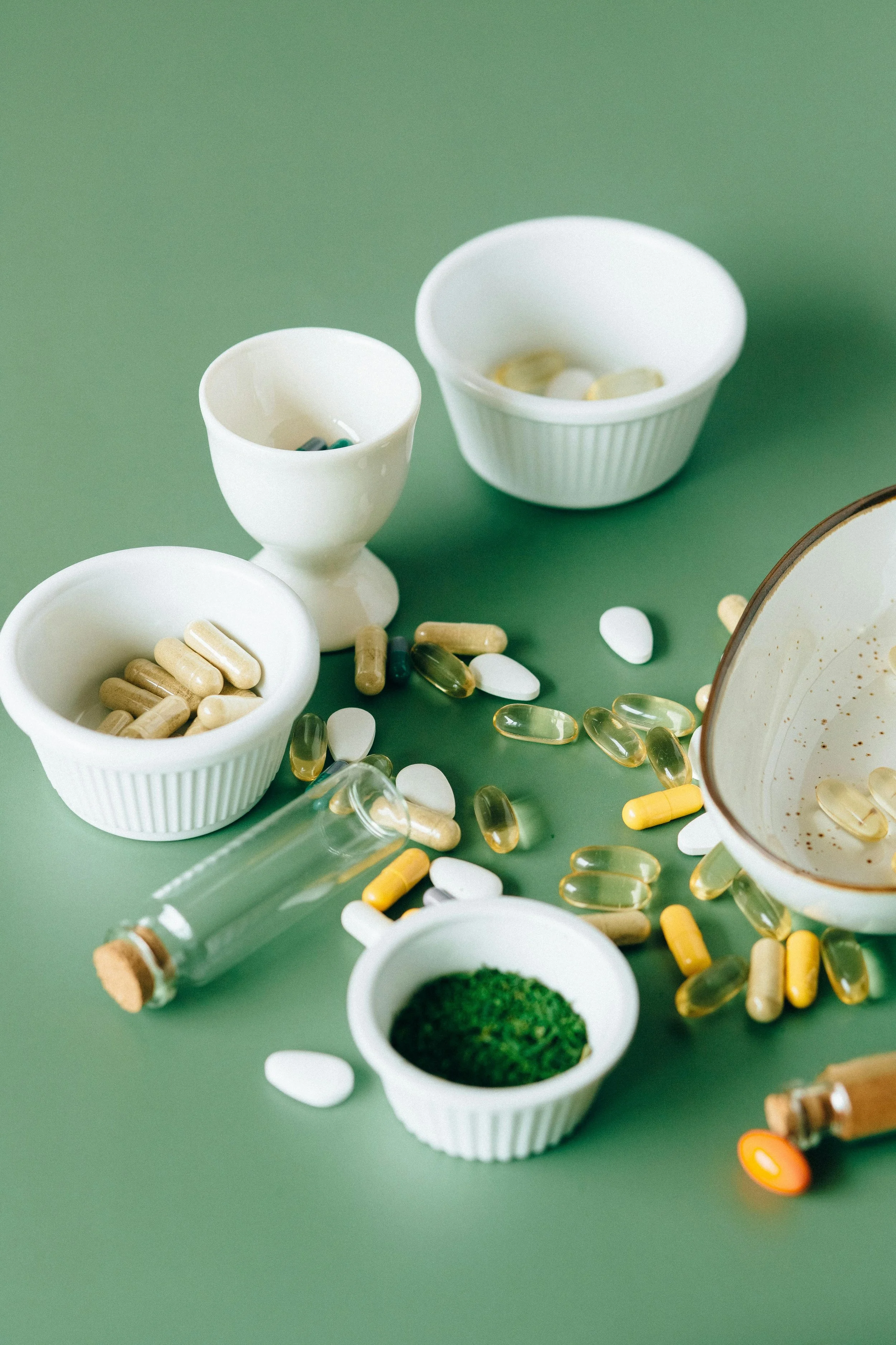 Various pills and capsules scattered on a green surface, with some in white bowls and small bottles.