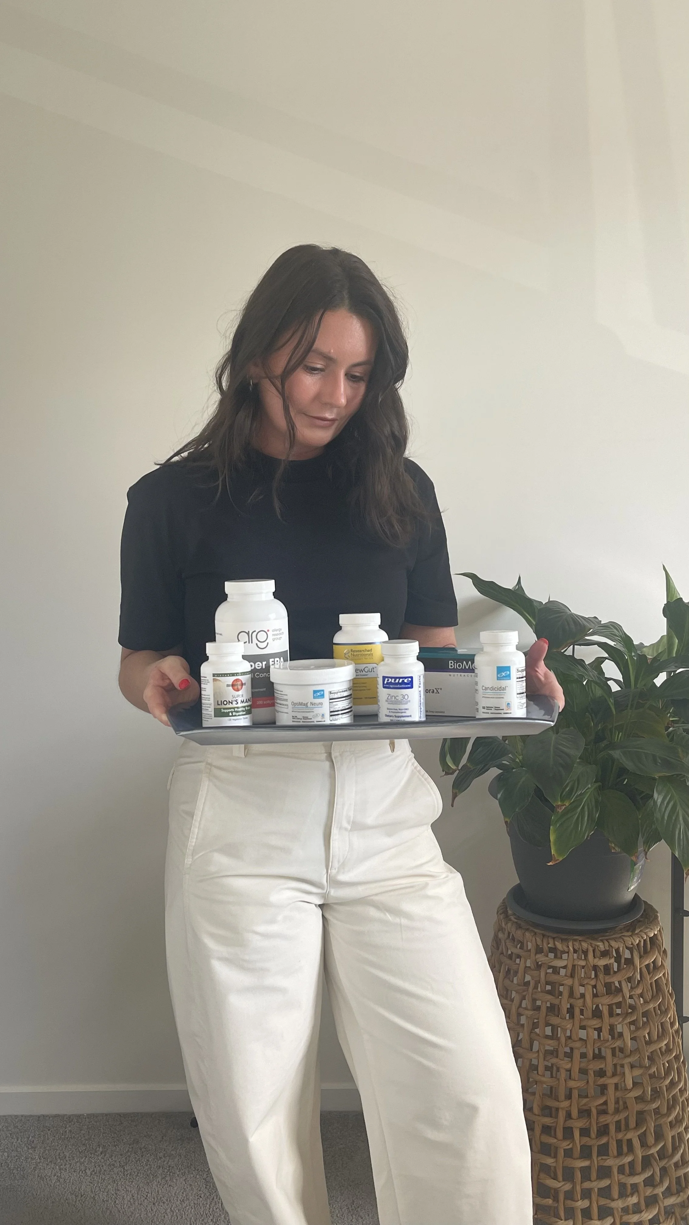 Woman holding a tray of various supplement bottles in a room with a potted plant.