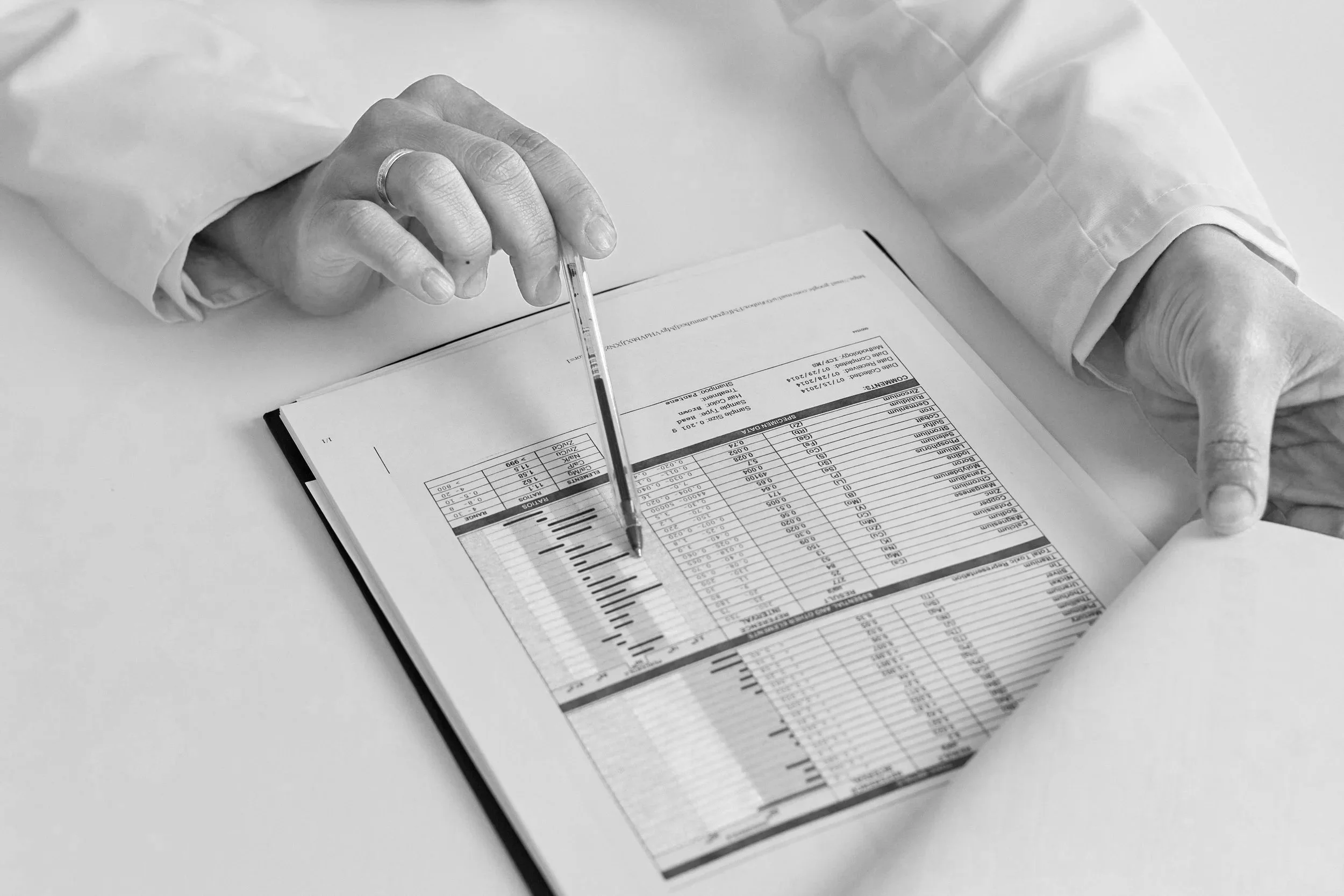A person in a lab coat pointing at a printed spreadsheet on a clipboard with a pen.
