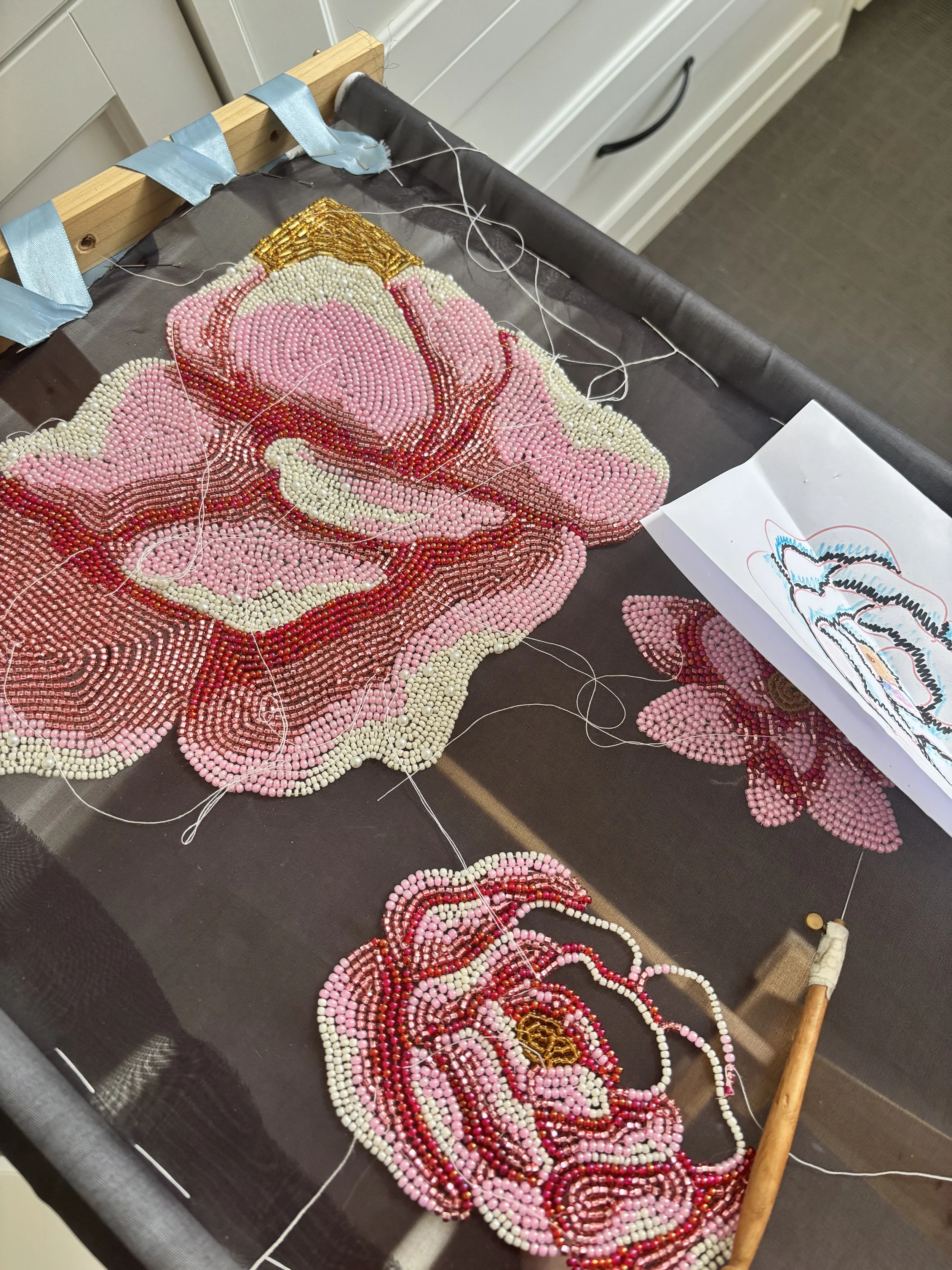 A beaded embroidery project in progress depicts a pink flower with white and red accents. The beads are sewn onto black fabric and are connected with white thread. A pattern sketch is visible on a piece of paper nearby, and a wooden tool with a threaded needle rests on the side, with a blue ribbon tied to the embroidery frame.