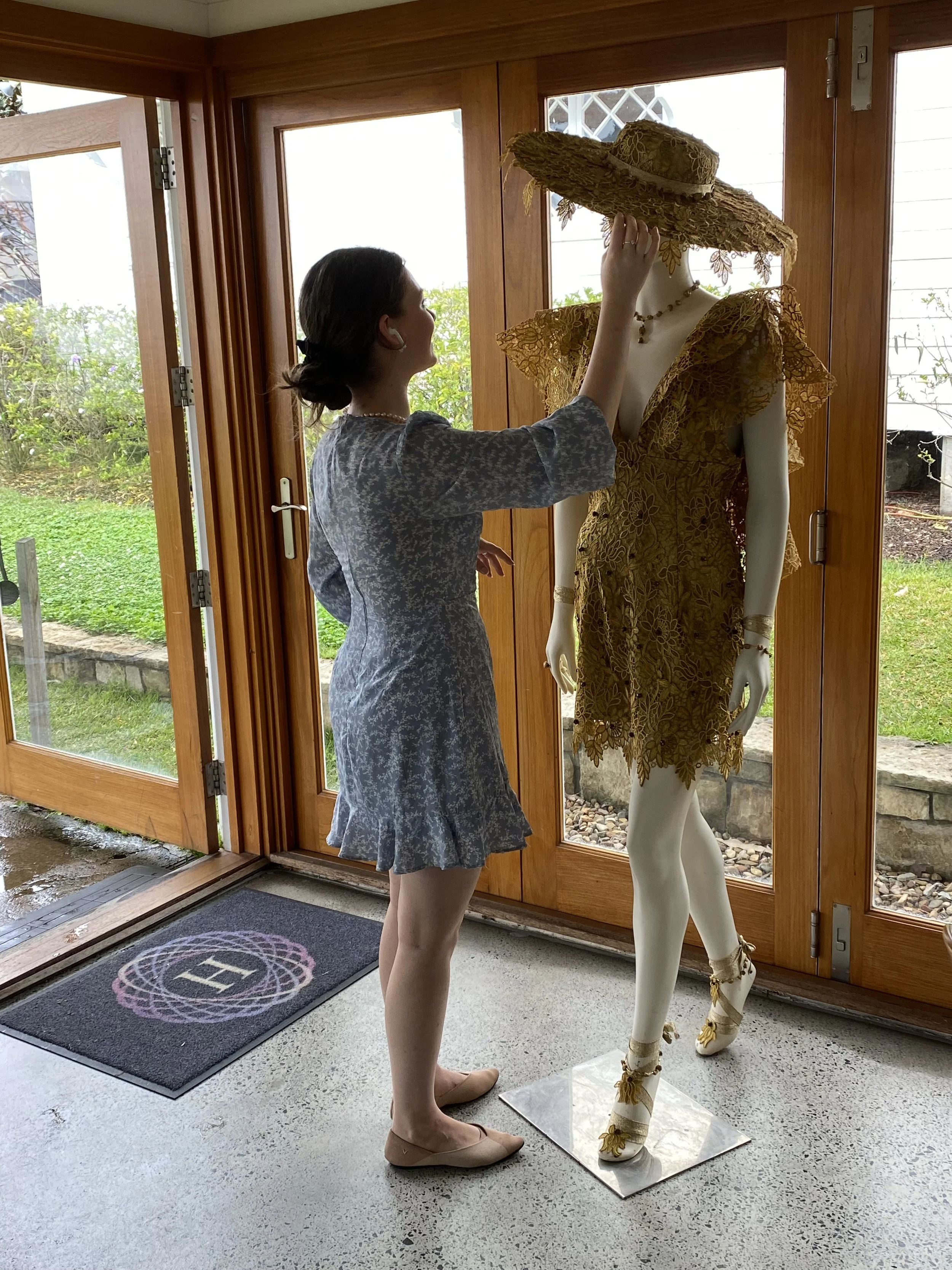 A woman in a dress and beige shoes is standing inside by a glass door. She is touching the brim of a mannequin's large hat. The mannequin is dressed in a yellow lace dress and floral ankle wrap shoes, standing on a metallic base. Outside, greenery and a garden are visible