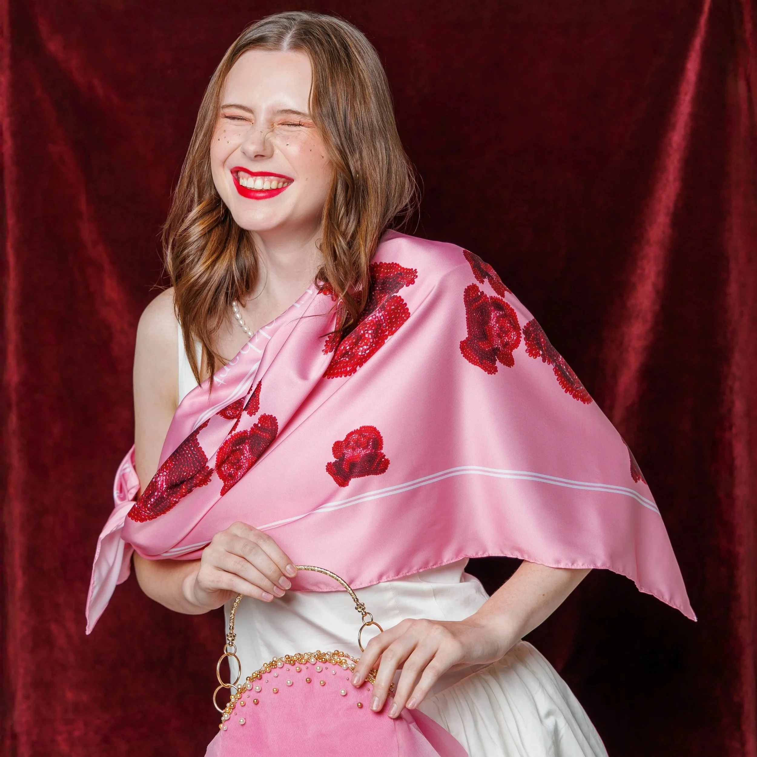A woman with brown hair, smiling with eyes closed, wearing a pink silk dress with red floral embroidery, holding a pink beaded handbag, standing in front of a red curtain.