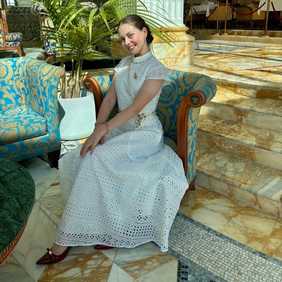 A young woman wearing a white eyelet dress sitting on a blue and gold patterned sofa in a luxurious hotel lobby with marble floors and stairs, green plants, and elegant furniture.