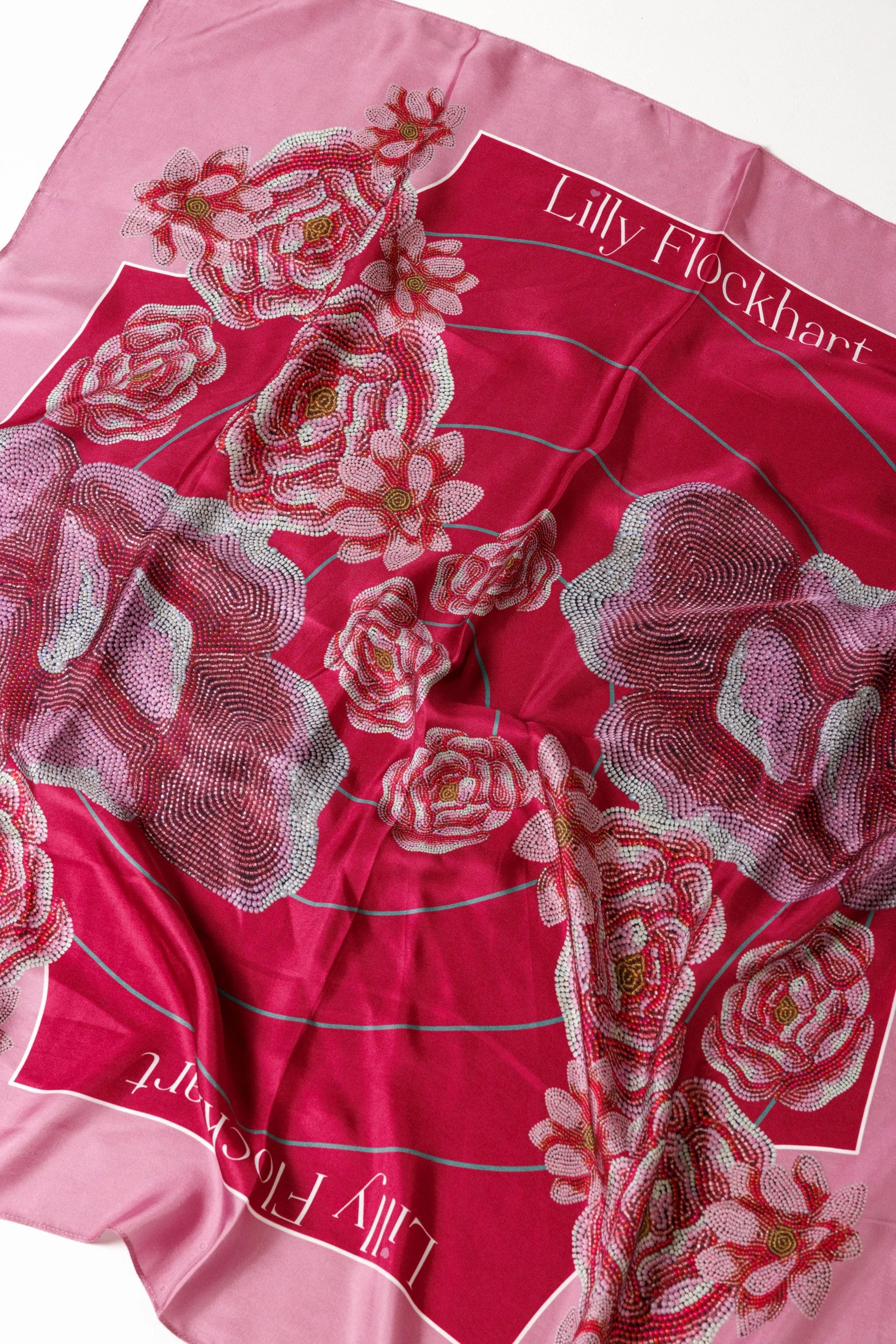 Pink silk scarf with a floral pattern of pink flowers with yellow centers and green stems, and a label that reads 'Lilly Flockhart' in white text on a pink background.
