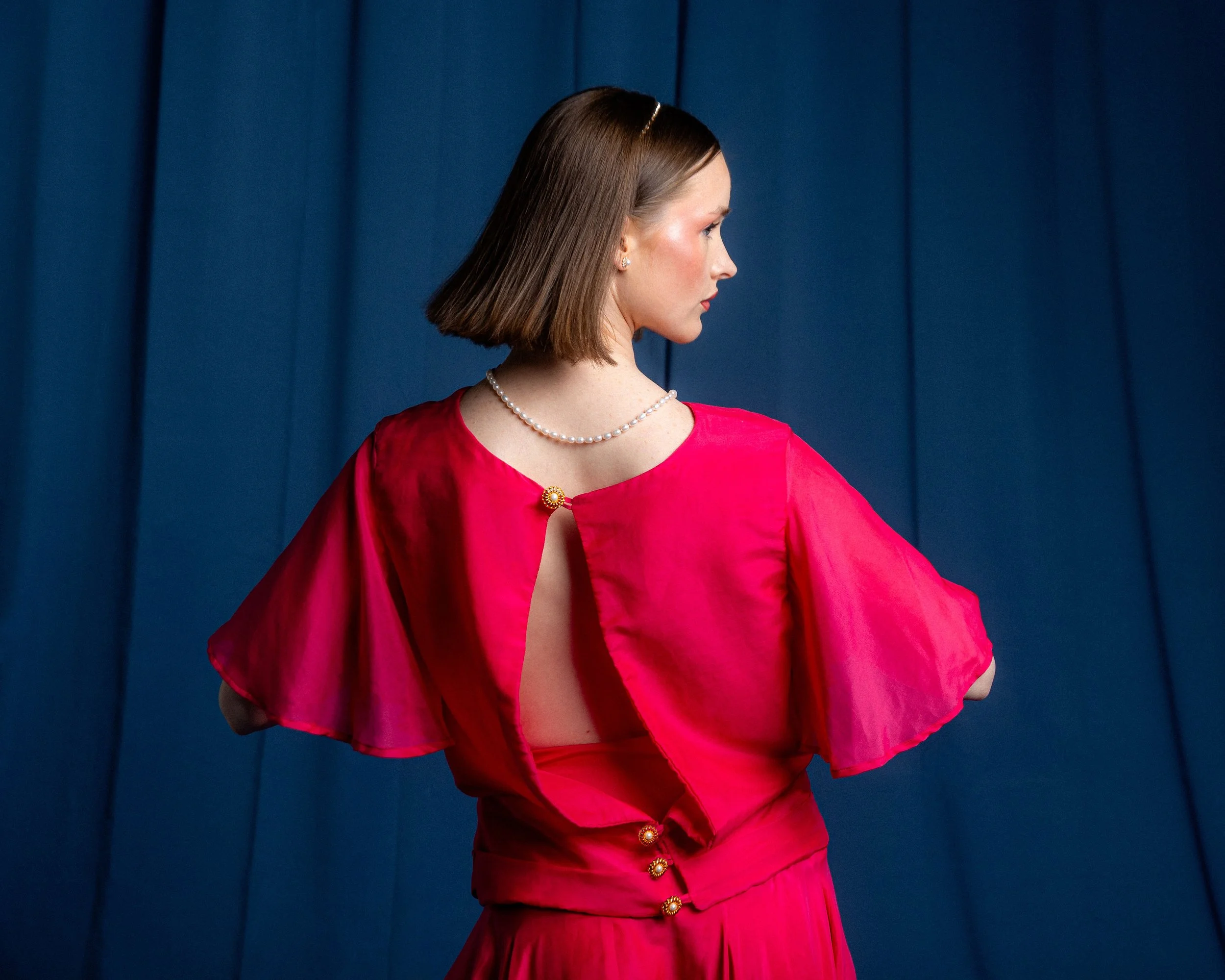 A woman with short brown hair in a bob style, wearing a pearl necklace, pearl earrings, and a bright red dress with decorative buttons, stands against a dark blue padded wall, looking to her right.