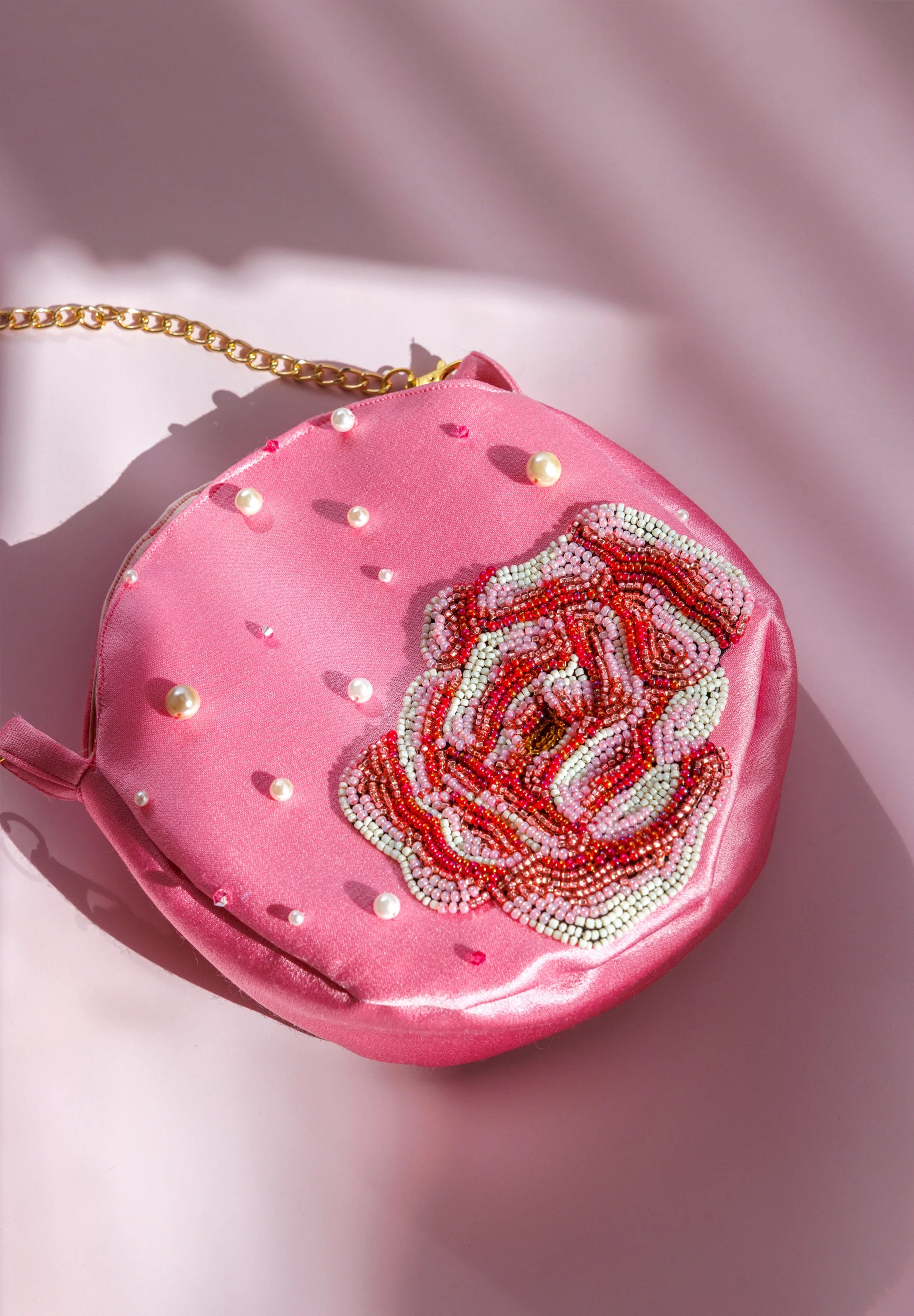 A pink satin purse adorned with small pearl accents and a detailed beaded embroidery of a colorful flower. The purse has a gold chain strap.