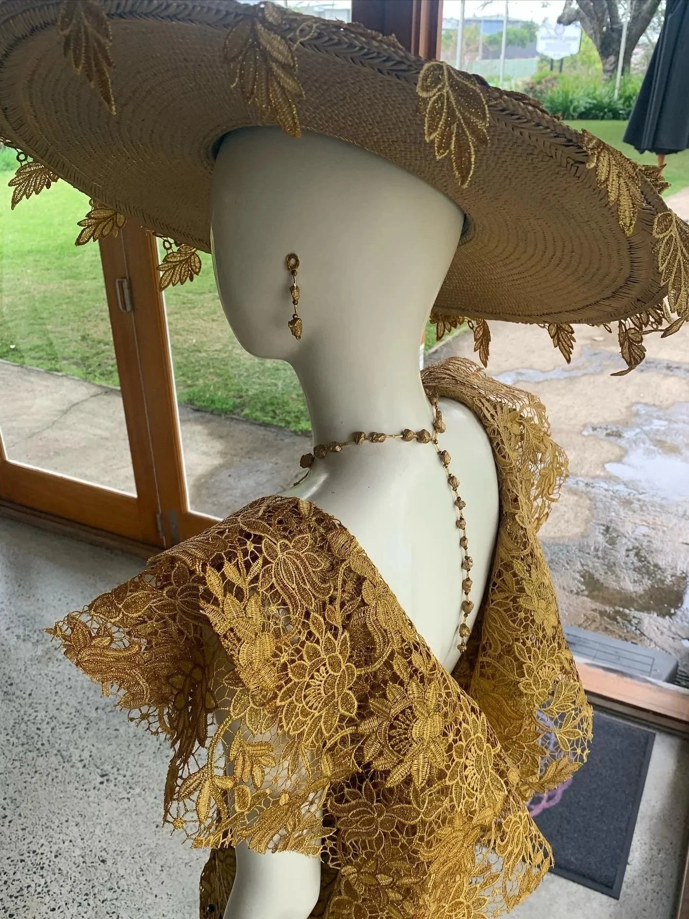 A mannequin dressed in a lace, gold-colored outfit with a large, woven hat decorated with gold embroidery and leaves. The mannequin is also wearing matching gold jewelry, including earrings, a necklace, and a long beaded chain. The background shows a grassy outdoor area visible through a large window.
