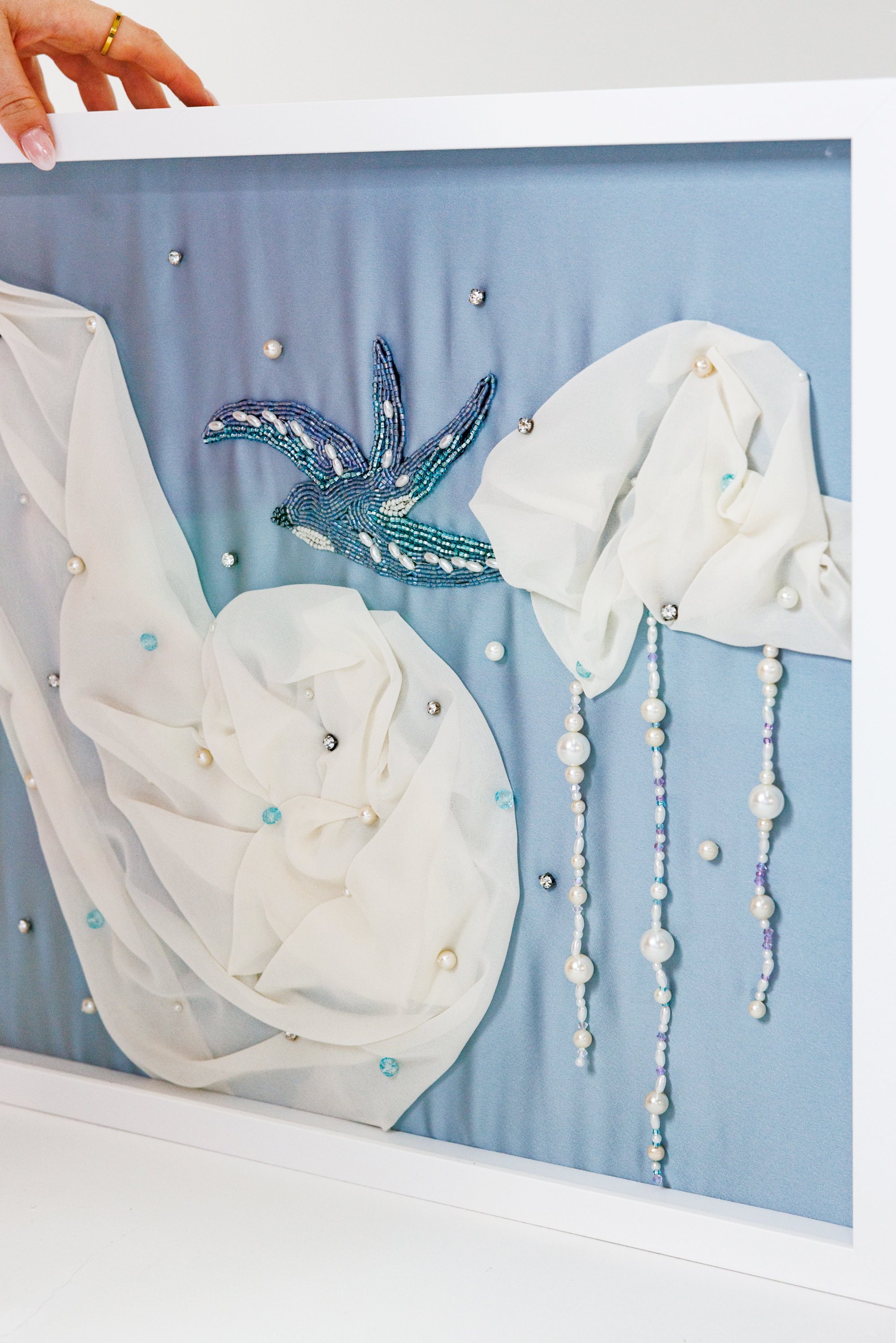 A framed decorative art piece featuring a beaded bird with blue and white beads, white fabric flowers, and hanging pearl and bead strands on a light blue background.