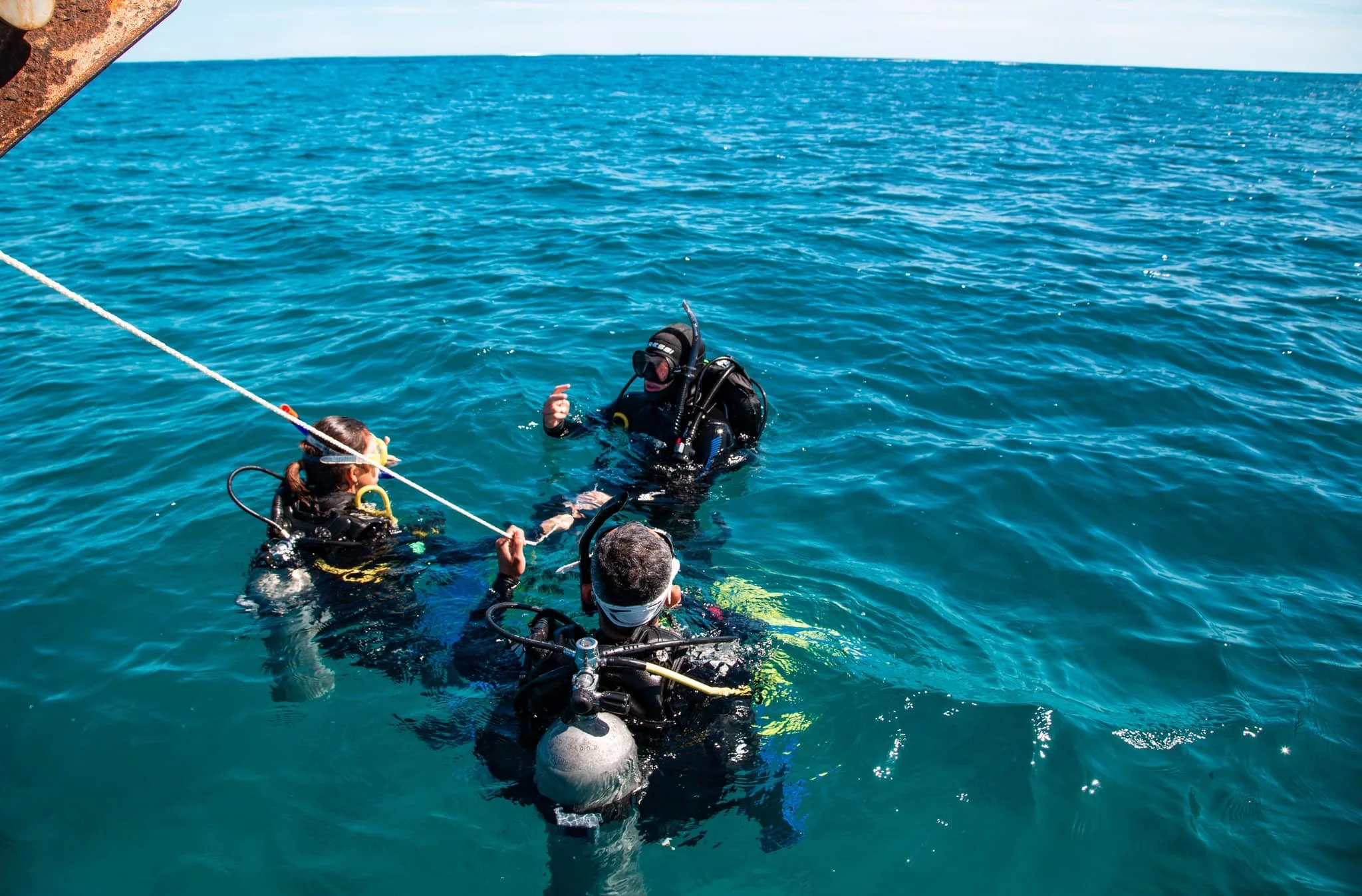 People scuba diving in the ocean, with two individuals in the foreground holding onto a boat's rope, wearing wetsuits, masks, and breathing equipment, surrounded by blue water and a clear sky.