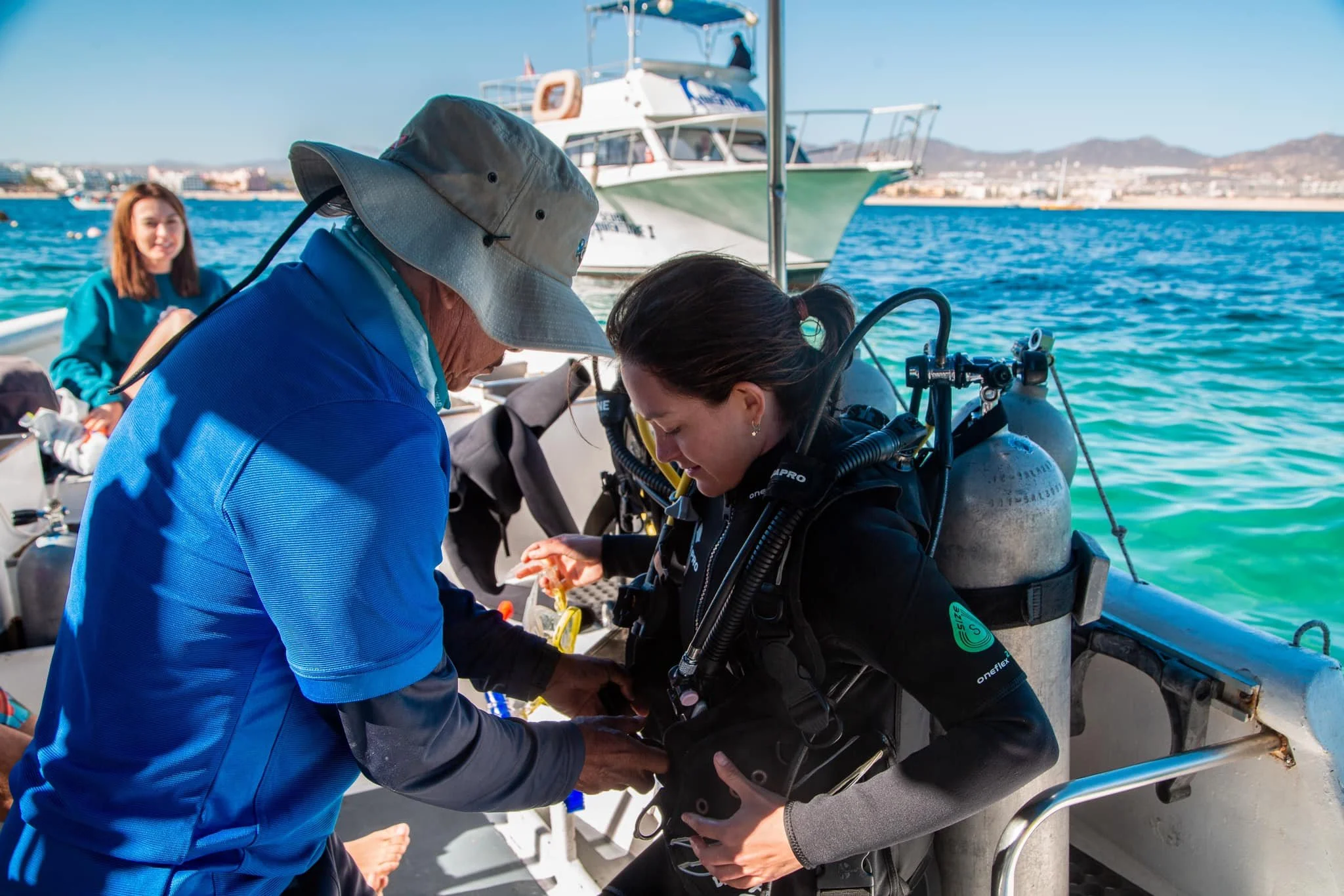People preparing scuba diving equipment on a boat near the water with boats and cityscape in the background.