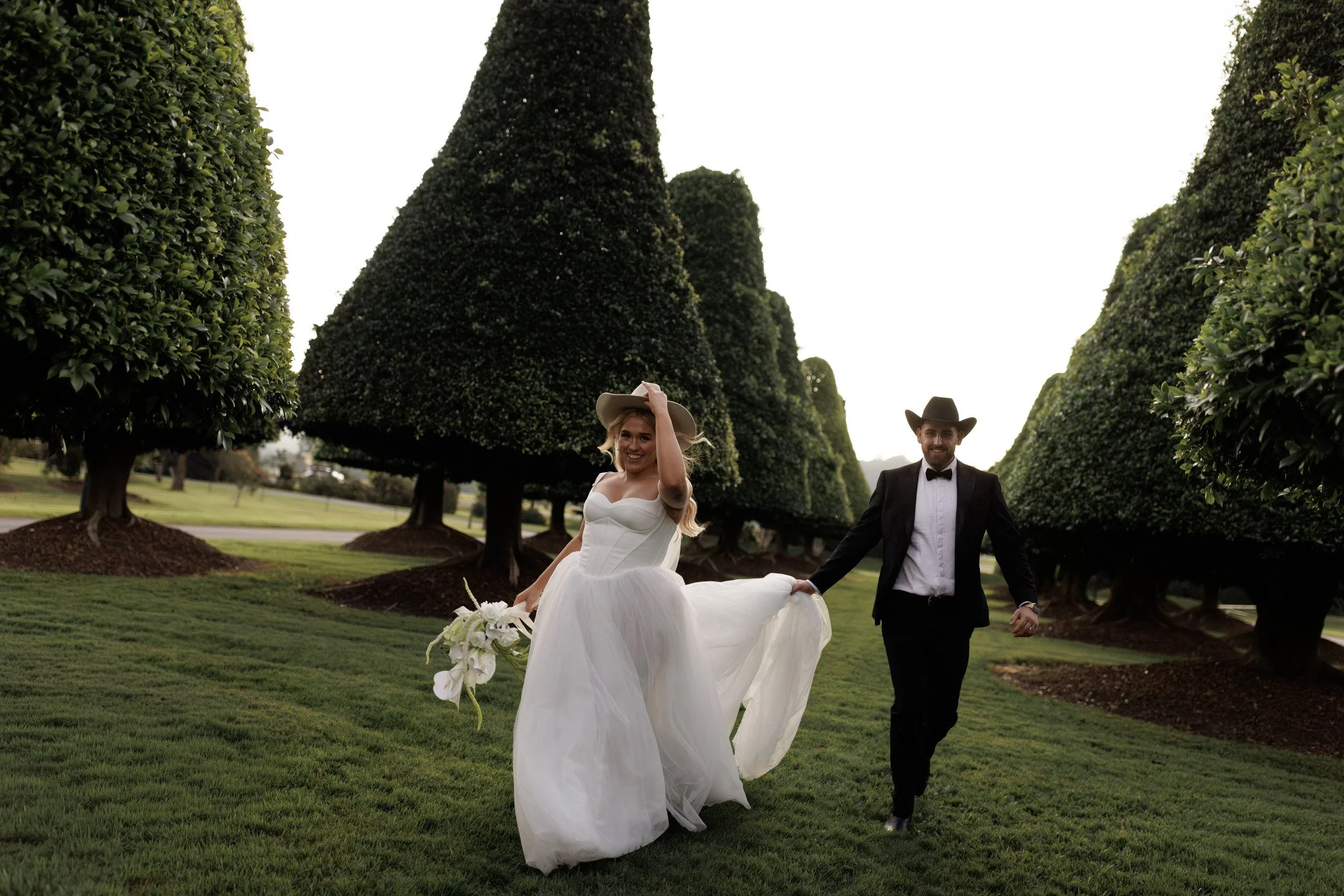A bride and groom holding hands walking outdoors among tall, sculpted green bushes on a sunny day.