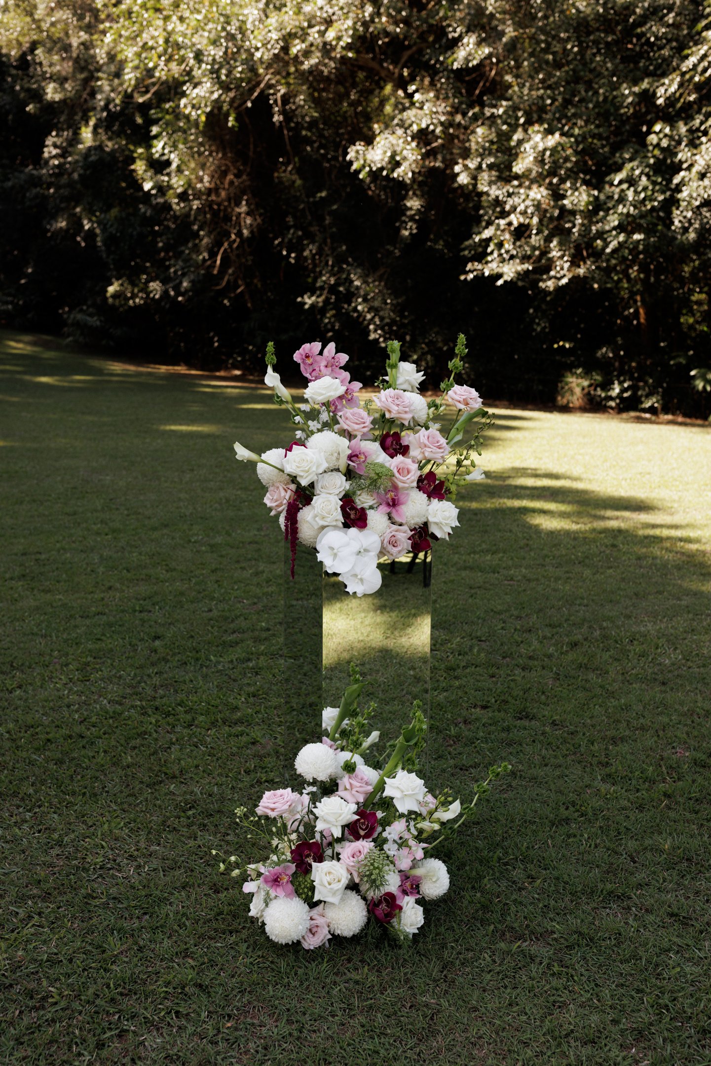 A tall, narrow floral arrangement featuring pink and white roses, purple orchids, and white pom-pom flowers, set outdoors on a grassy area with trees in the background.