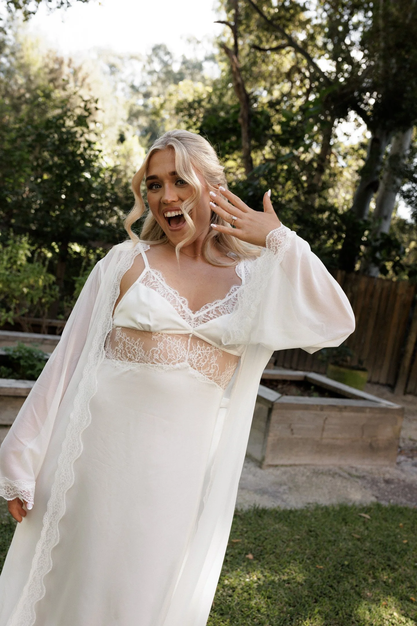 A woman in a white dress with lace details, smiling and showing her engagement ring, outdoors with trees and a wooden fence in the background.