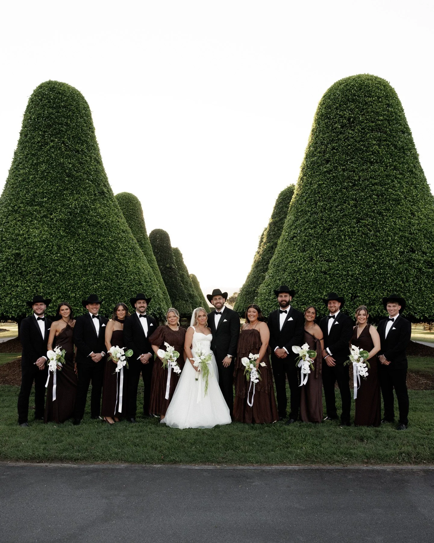 A group of people dressed in formal attire, including a bride in a white wedding gown, standing outdoors on grass in front of tall, neatly trimmed green trees, possibly at a wedding or special occasion.