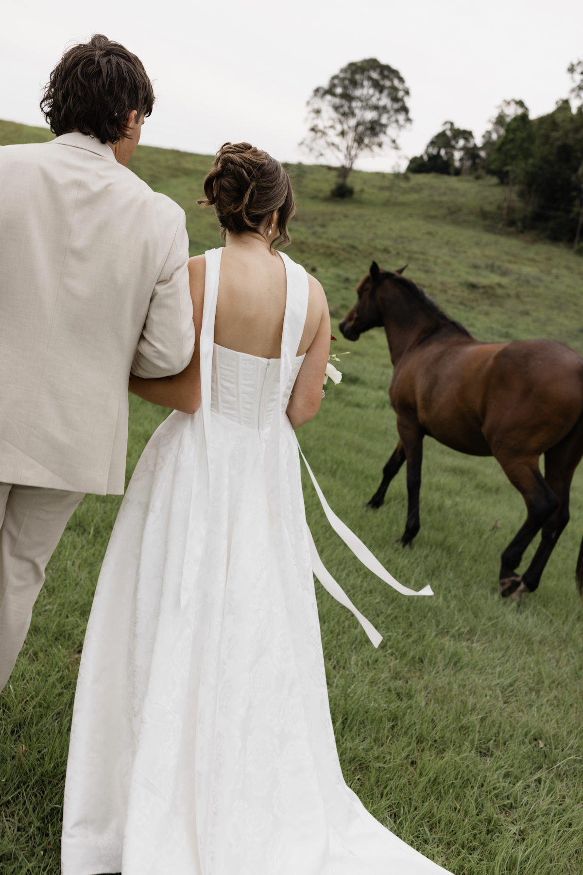 A bride and groom walk together in a grassy field, with the bride holding a flower bouquet, and a brown horse nearby, surrounded by trees and rolling green hills.