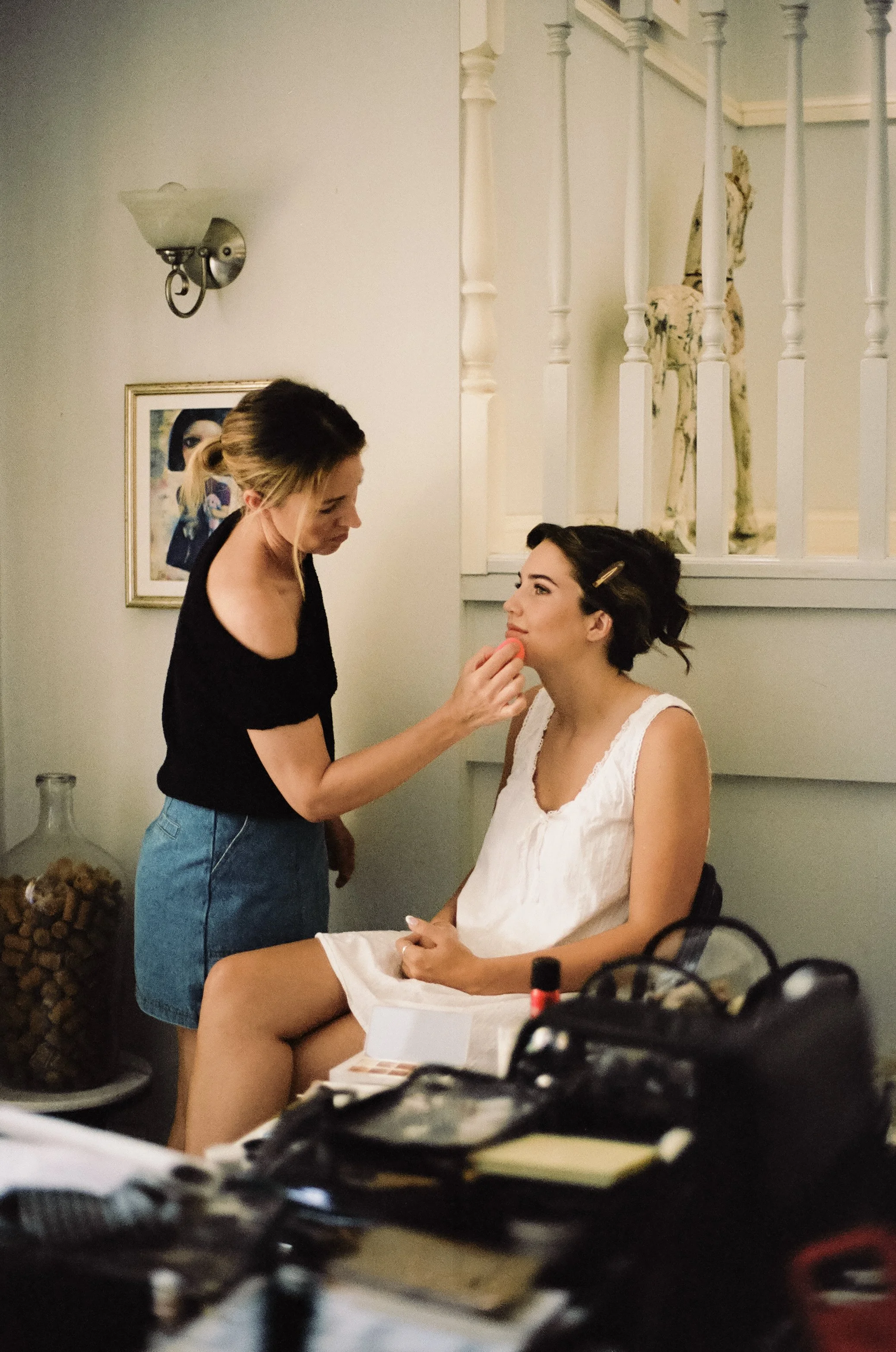 A woman sitting on a chair with a white dress is having makeup applied by another woman in a black top in a white room.