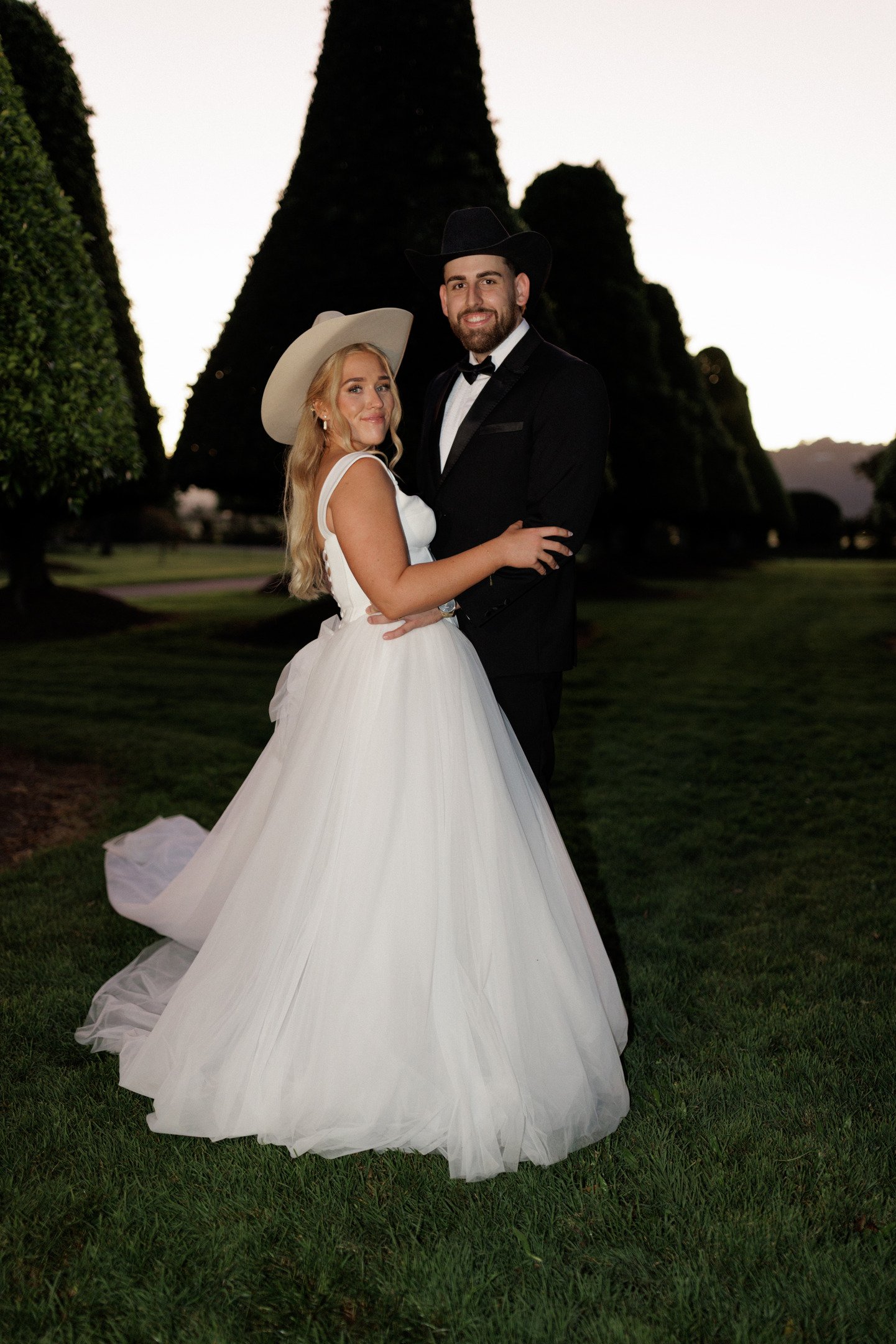A bride and groom standing outdoors at sunset, with tall trees in the background. The bride wears a white wedding gown and large hat, and the groom wears a black suit with a bow tie and cowboy hat. They are smiling and holding each other.