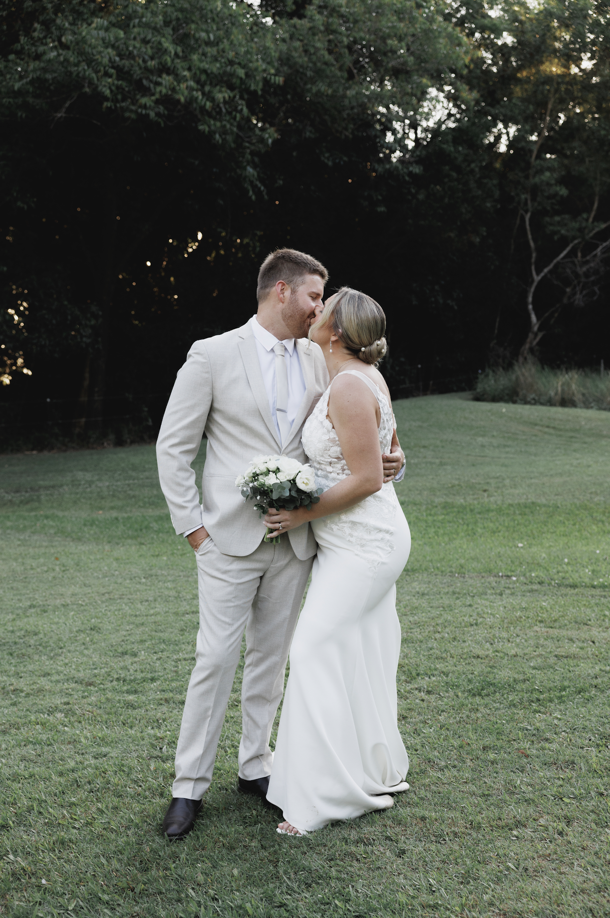 A newlywed couple sharing a kiss outdoors on a grassy field with trees in the background, the bride holding a bouquet of white roses.