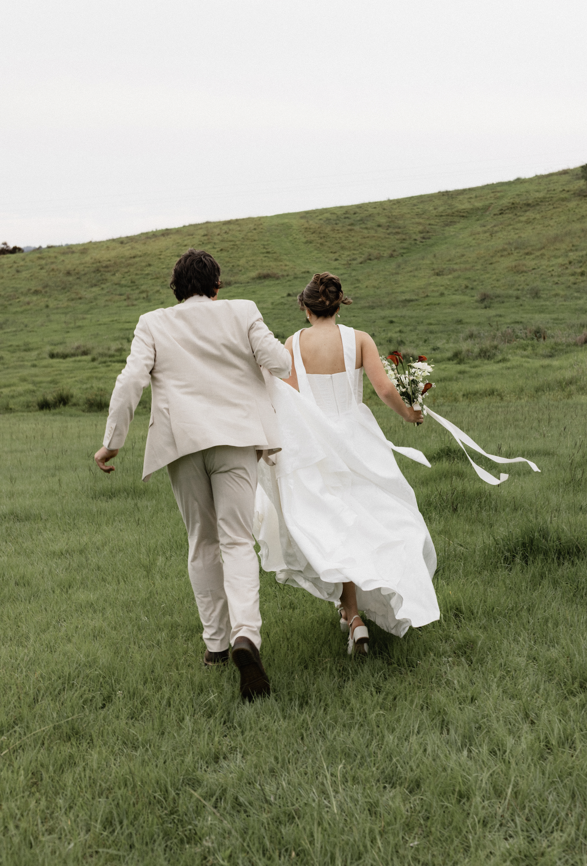 A couple, dressed in wedding attire, running through a grassy field with a green hill in the background, the bride holding a bouquet of flowers.