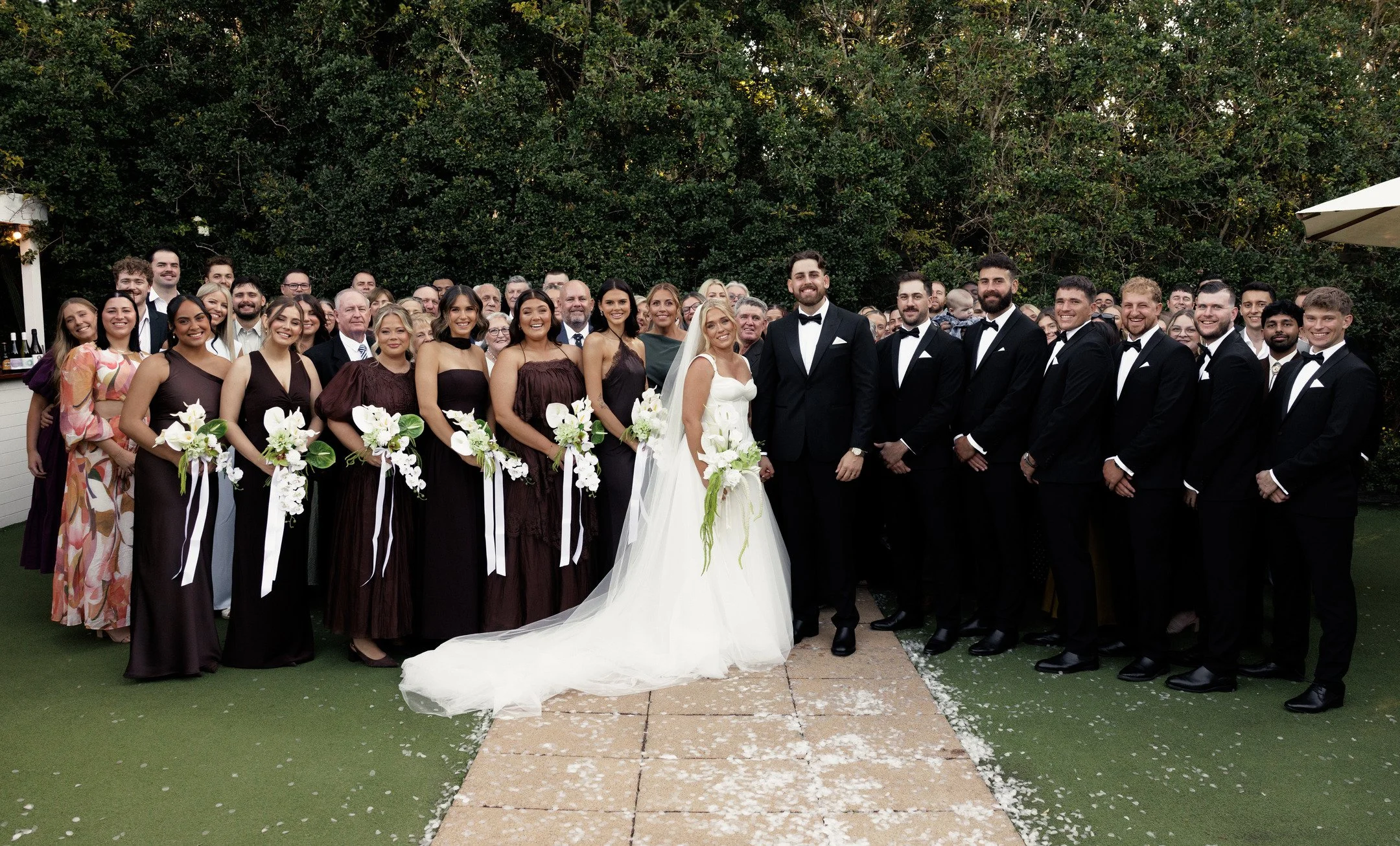 A large group of wedding guests gathered outdoors, with a bride and groom at the front center, dressed in formal attire, posing for a group photo in front of a green leafy background.