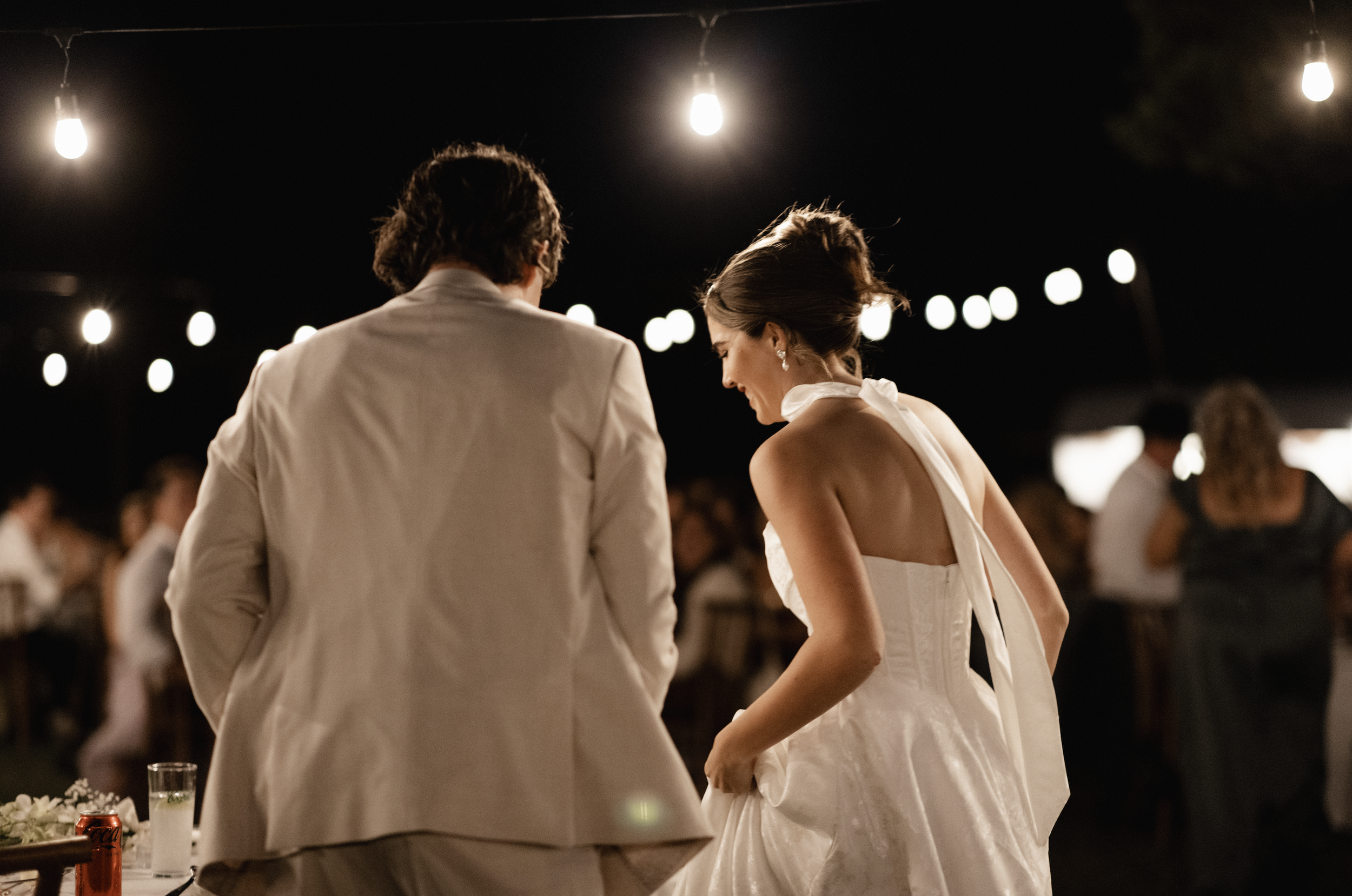 A bride and groom at their wedding reception under string lights at night, with guests in the background.