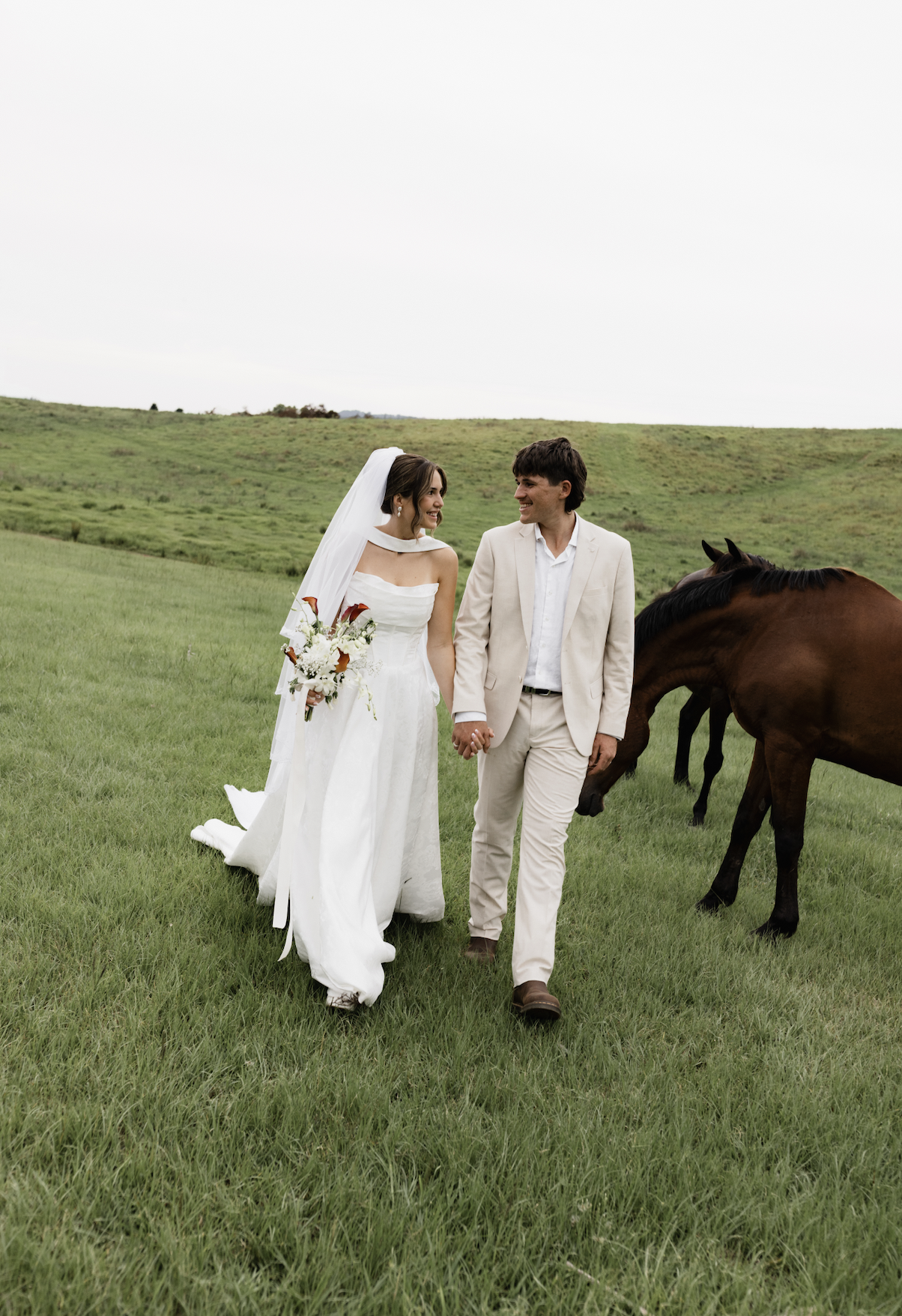 A bride and groom walking hand in hand in a green field, with brown horses grazing nearby, on a cloudy day.