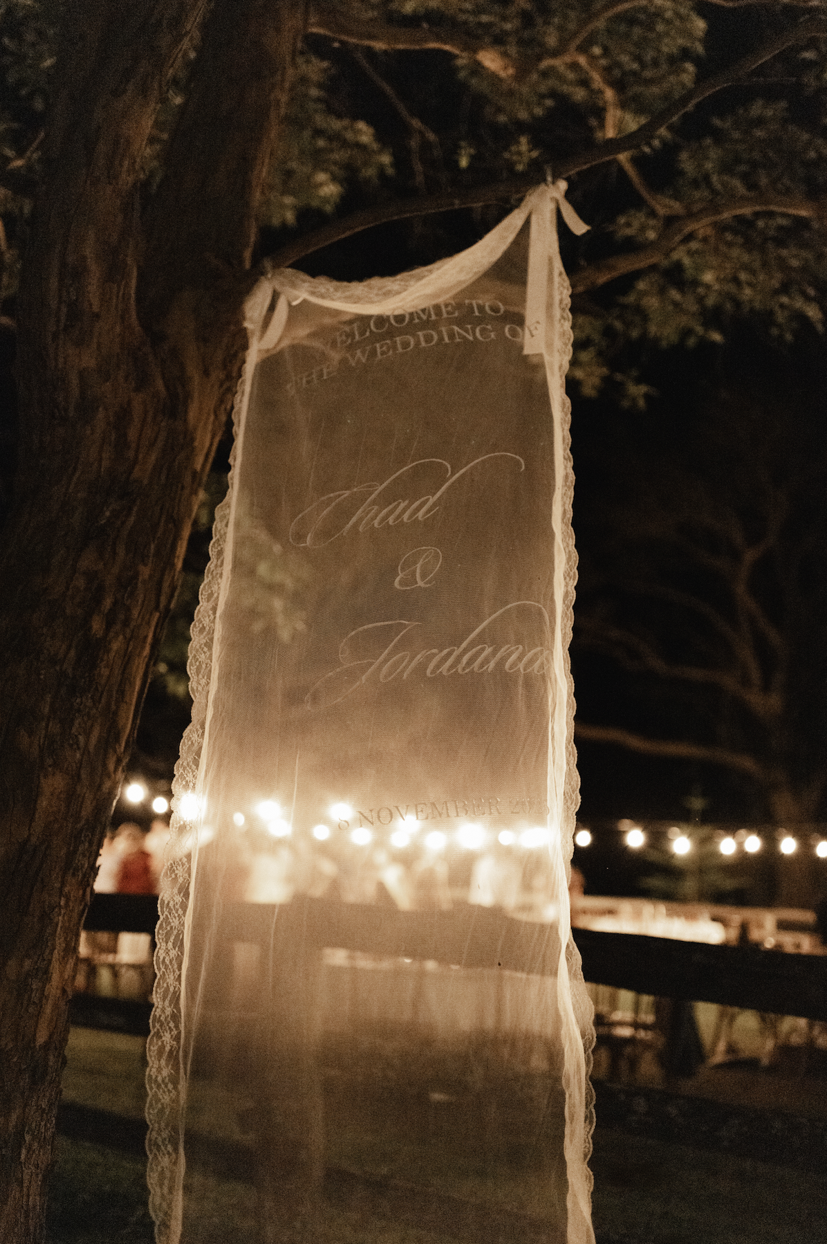 Decorative veil or banner hanging from a tree at night, with string lights in the background, announcing a wedding ceremony for Chad and Jordana.
