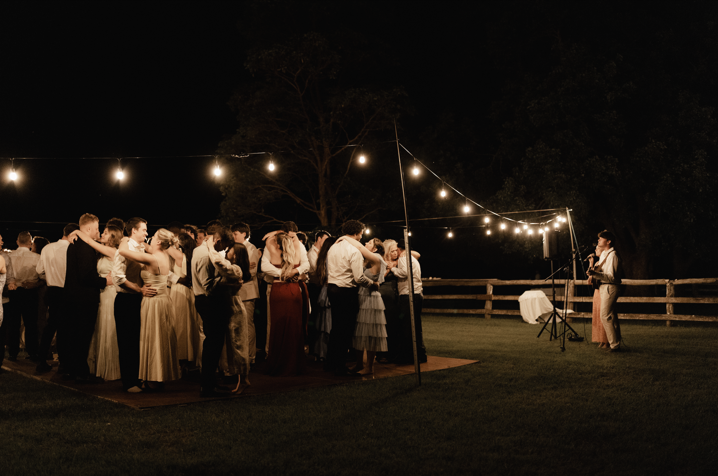 Guests dancing at an outdoor wedding reception at night, illuminated by string lights with trees and a wooden fence in the background.