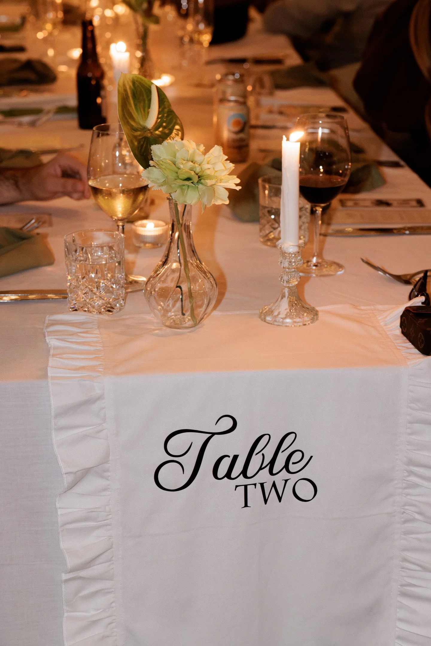 A decorated dining table with candles, glasses of white and red wine, a flower arrangement in a vase, and greenery. The table has a white tablecloth with ruffled edges and a sign indicating 'Table Two' in elegant script.