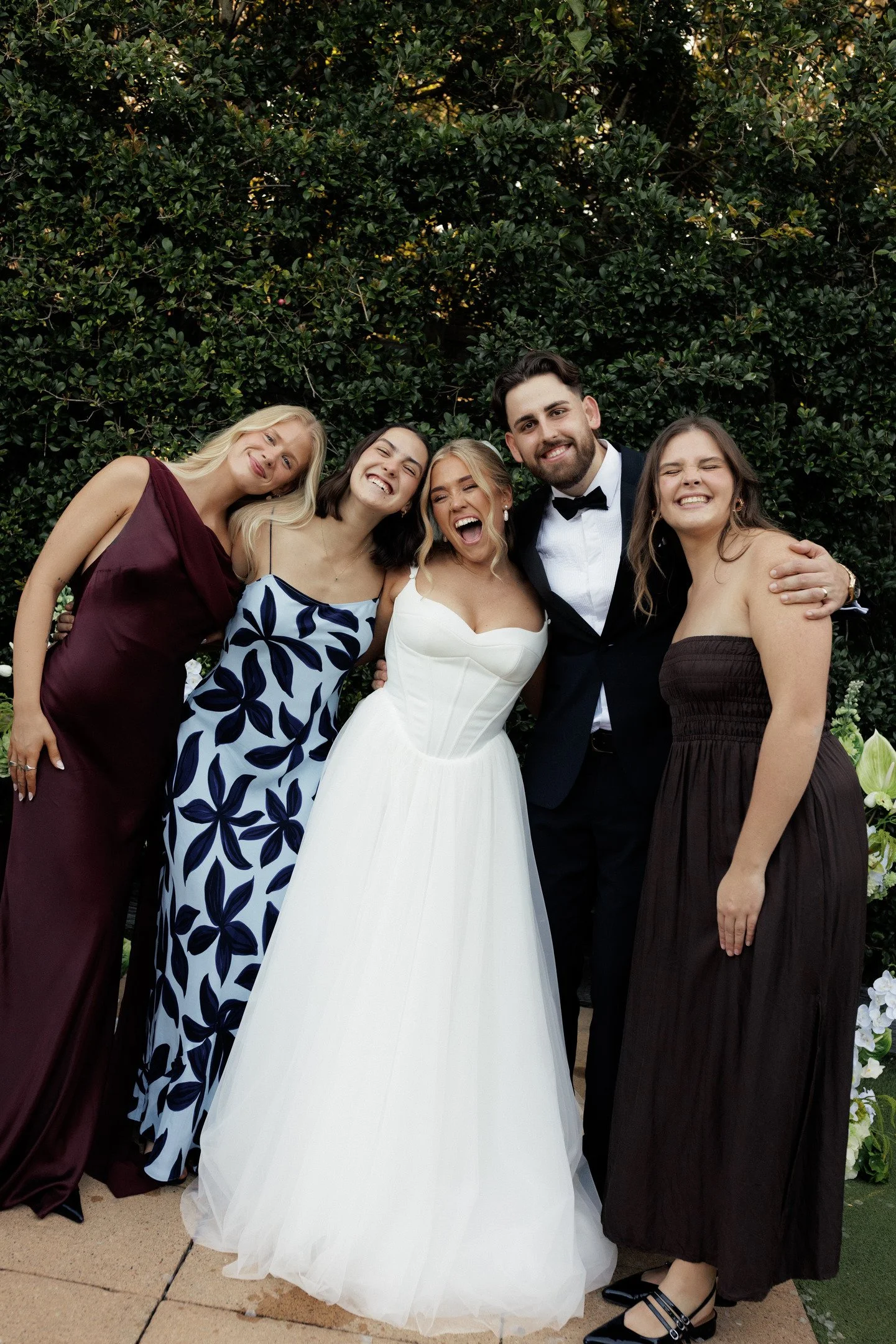 Group of five young adults dressed in formal attire, smiling and posing outdoors in front of a hedge, during a wedding celebration.