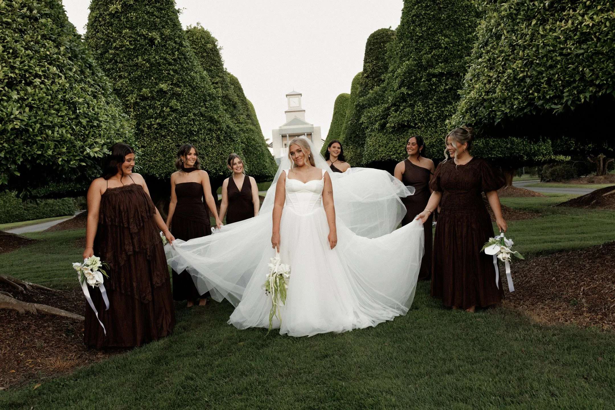 A bride in a white wedding gown and veil walking on a grassy path, surrounded by six women in brown dresses holding white flowers, with neatly trimmed tall hedges and a small building with a clock tower in the background.