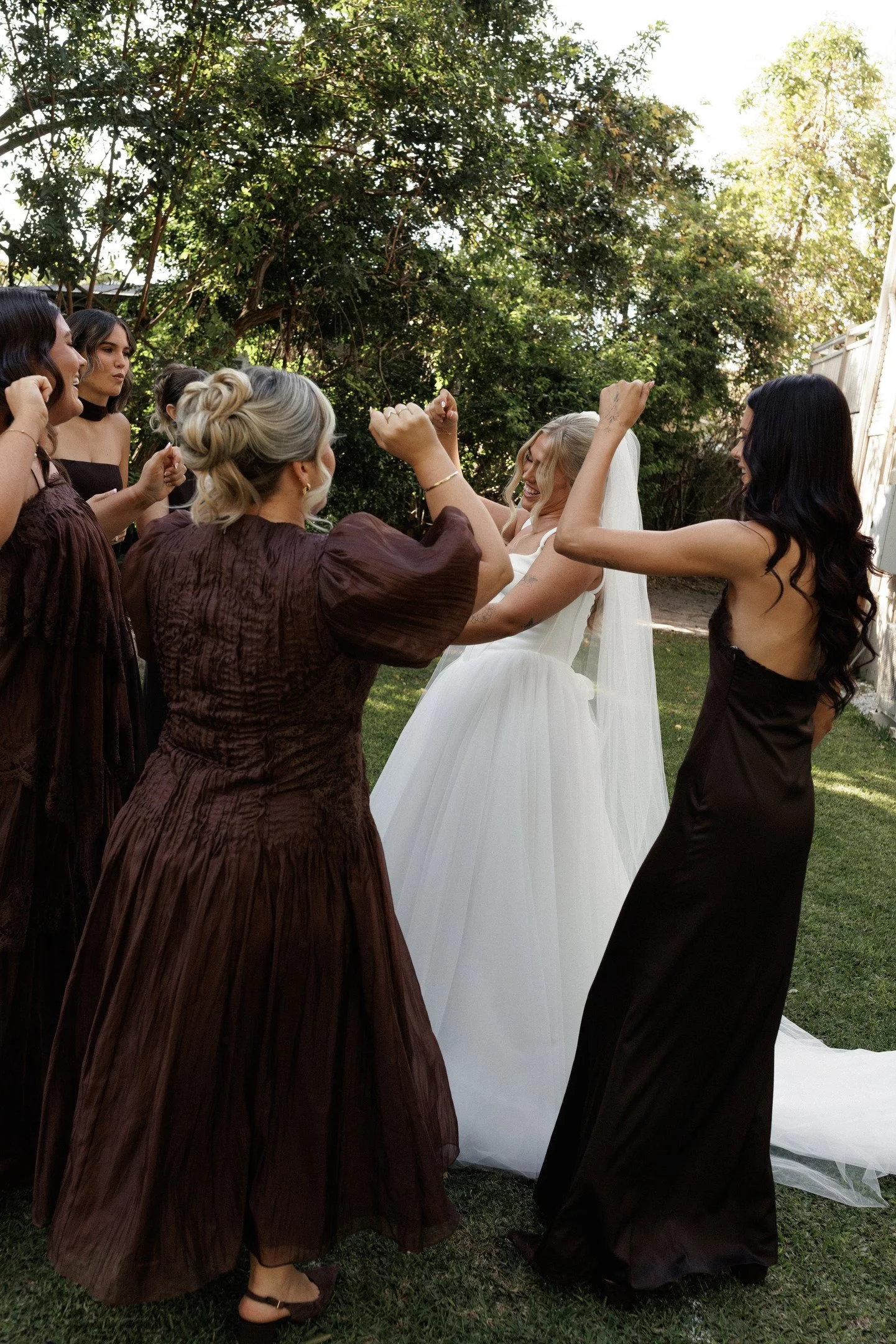 A group of women, including a bride in a white wedding dress and her bridesmaids in brown dresses, are celebrating outdoors with trees in the background, raising their arms and smiling.