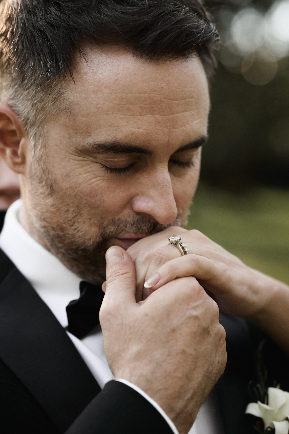 A man in a tuxedo kissing a woman on the hand, with his eyes closed. The man has dark hair and a beard, and is wearing a ring with a large gemstone on his finger. The background is blurred with greenery.