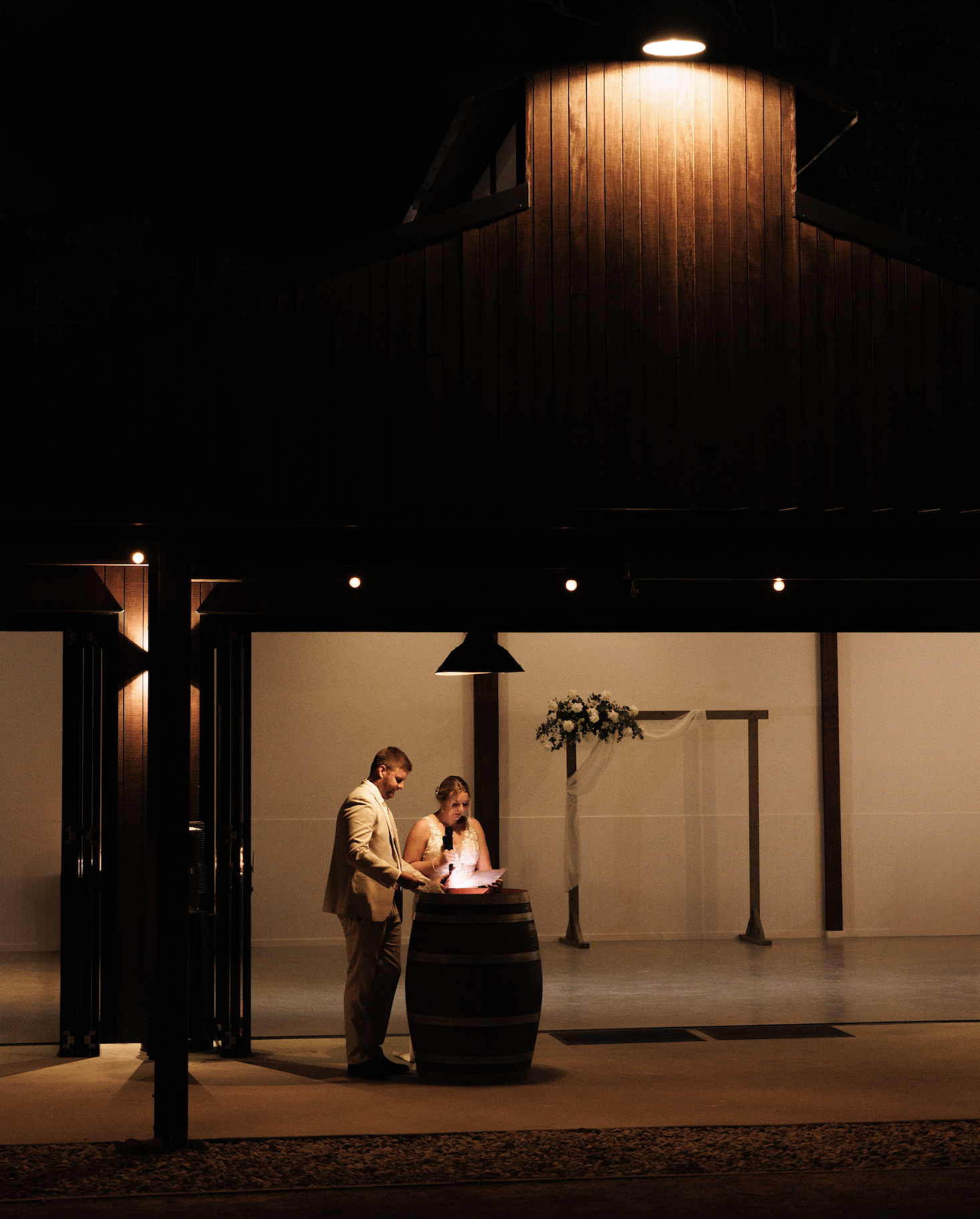 A couple dressed in wedding attire stands at a table with a microphone and papers, in a dimly lit indoor venue with a wooden wall and floral decoration in the background, possibly during a wedding reception or ceremony.