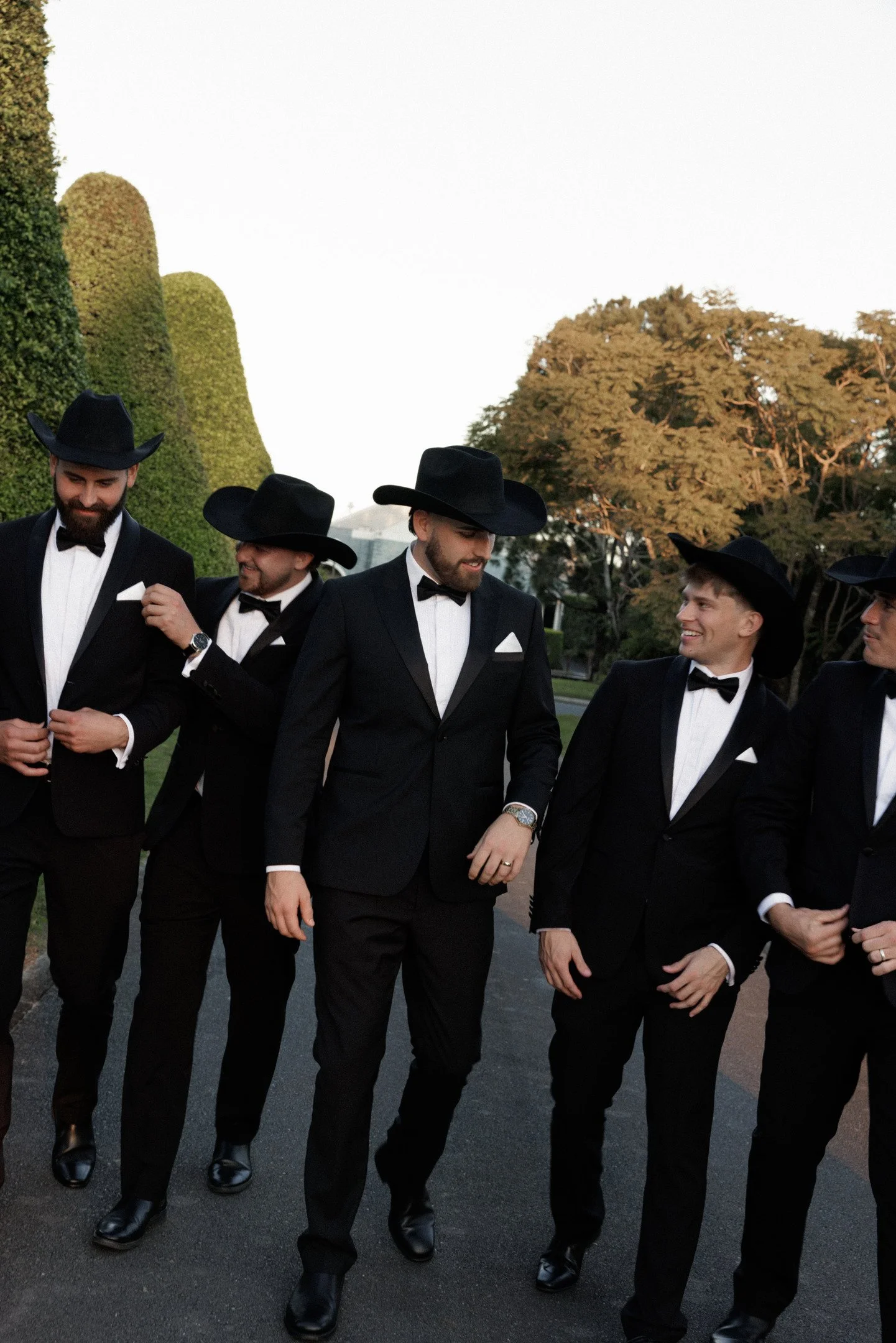Group of men in black Western suits, bow ties, and black cowboy hats walking together outdoors during the daytime, with trees and clear sky in the background.