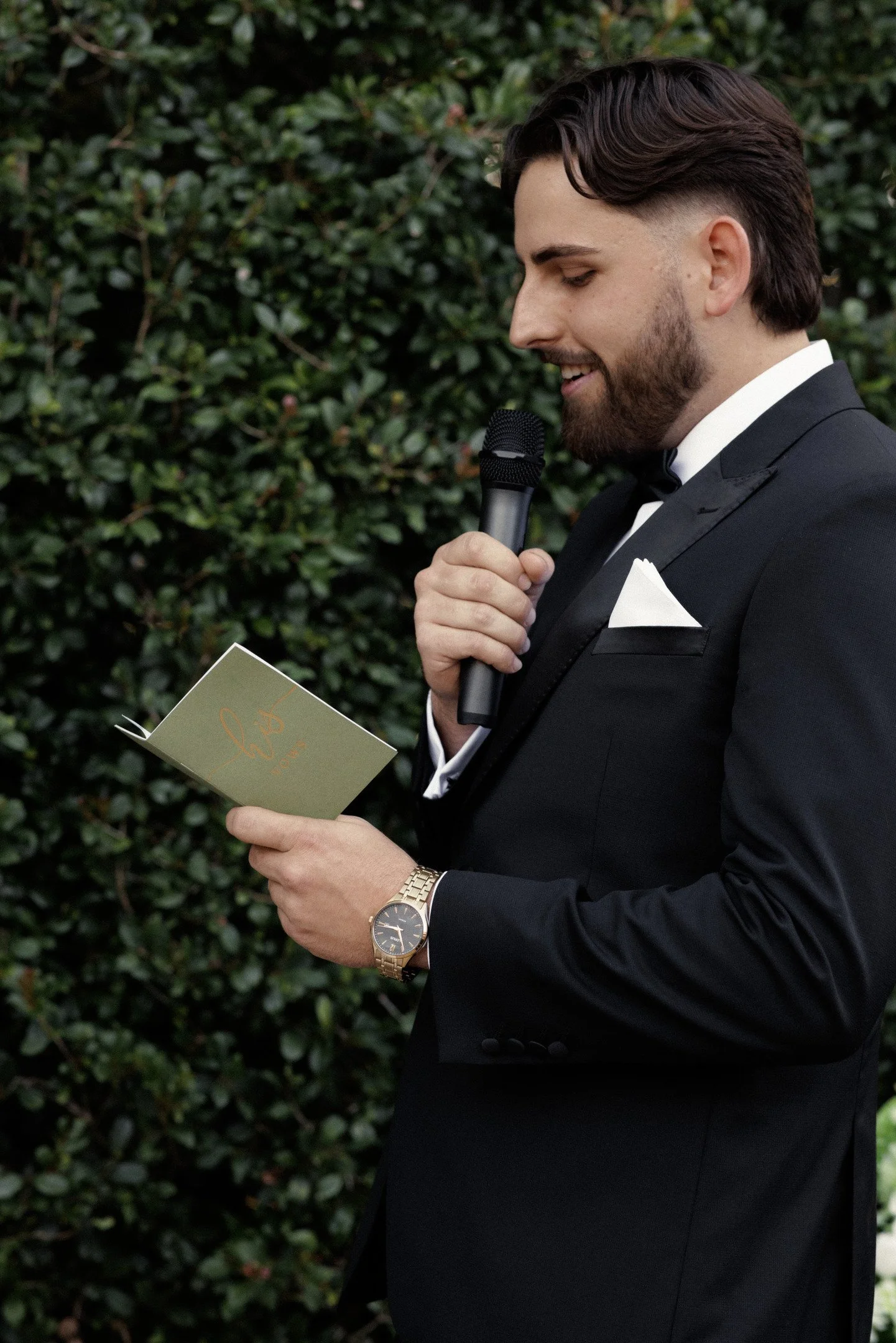 A man in a tuxedo holding a microphone and reading from a card during an outdoor event, with a background of green shrubbery.