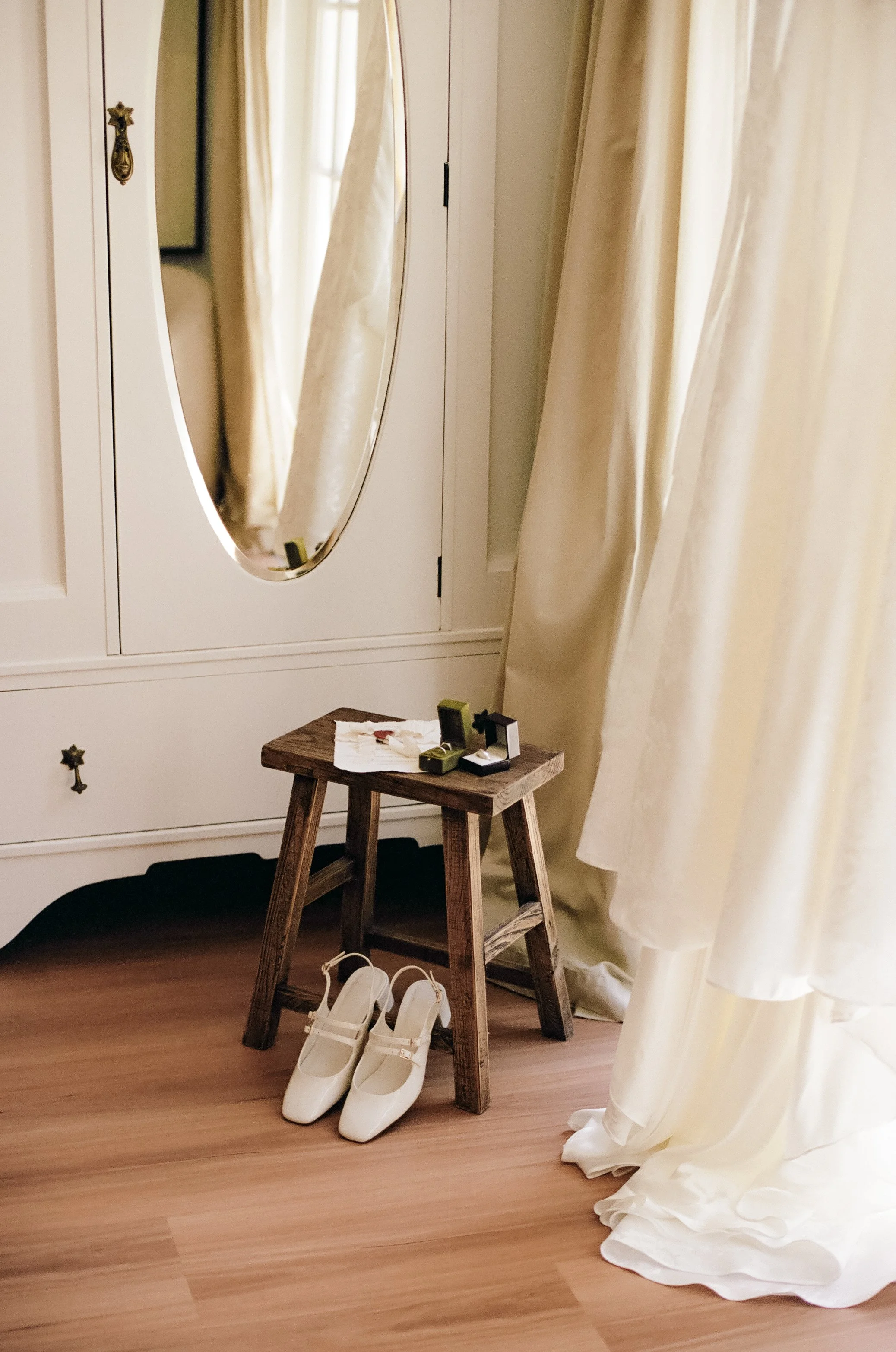 White bridal shoes on the floor, a small wooden stool with jewelry boxes and a napkin on top, white curtains, a large white wardrobe with an oval mirror reflecting the room.